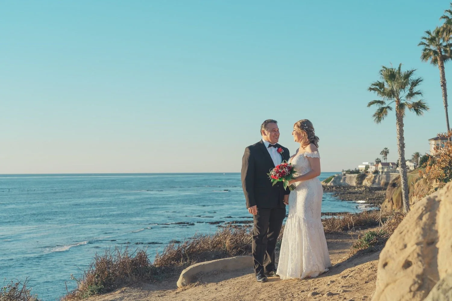A bride and groom standing on a beach, holding hands and smiling at each other. The bride is wearing a white wedding gown and holding a bouquet of red and pink flowers. The groom is dressed in a black tuxedo. Tall palm trees and beach houses are visi