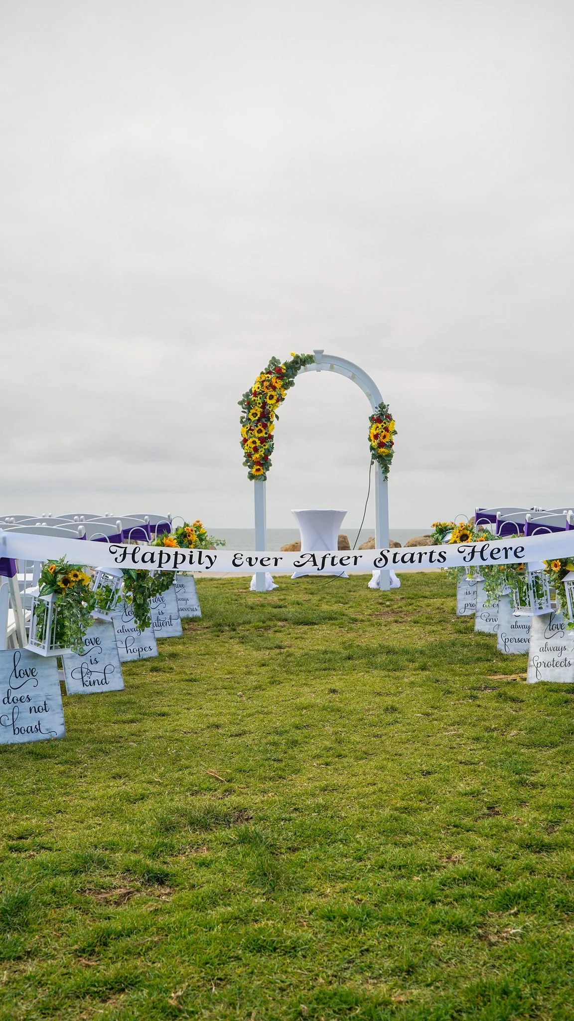 Outdoor wedding ceremony setup on a grassy area with an arch decorated with yellow and red flowers, chairs with purple sashes, and signs with inspirational quotes, under cloudy sky.