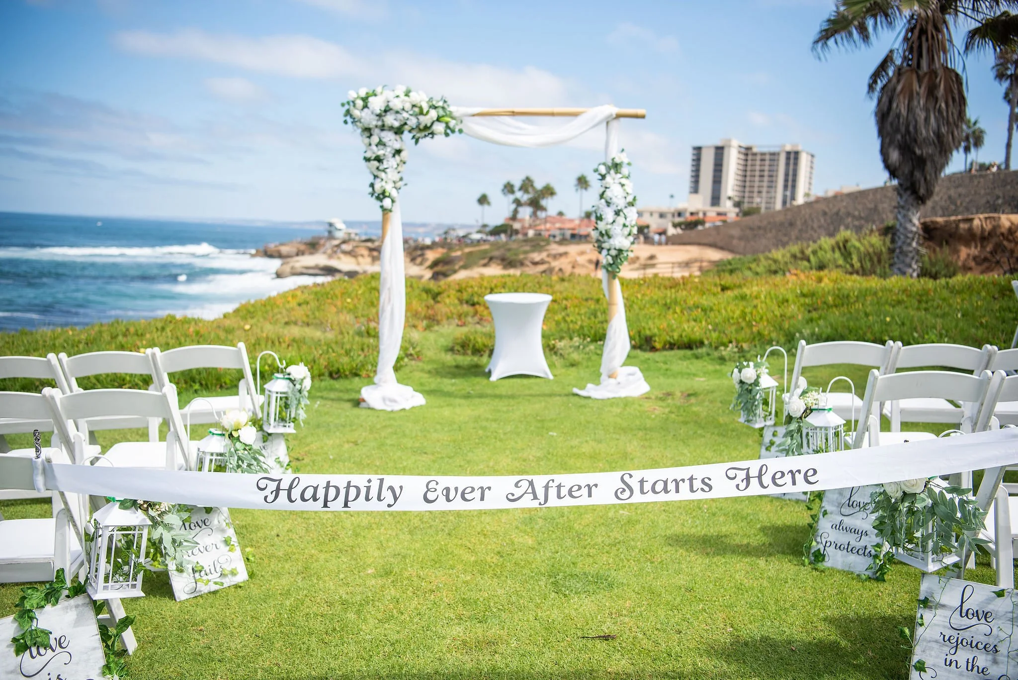 A wedding altar decorated with white flowers and drapery on a beachside lawn, with a scenic view of the ocean, palm trees, and buildings in the background.