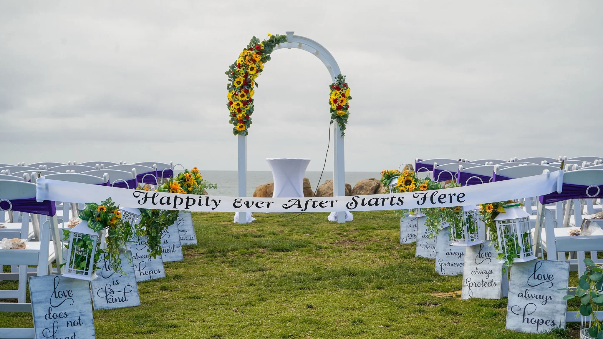 A decorated outdoor wedding ceremony setup with rows of white chairs with purple sashes, floral arrangements of sunflowers and greenery, and a white arch decorated with sunflowers and other flowers, with a beach and ocean in the background, and a whi