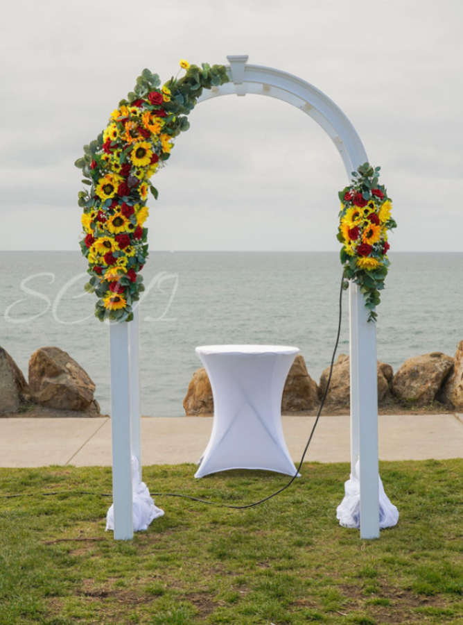 A white wedding arch decorated with yellow and red sunflowers and greenery, set up outdoors by the ocean with rocks in the background, and a small white table underneath.