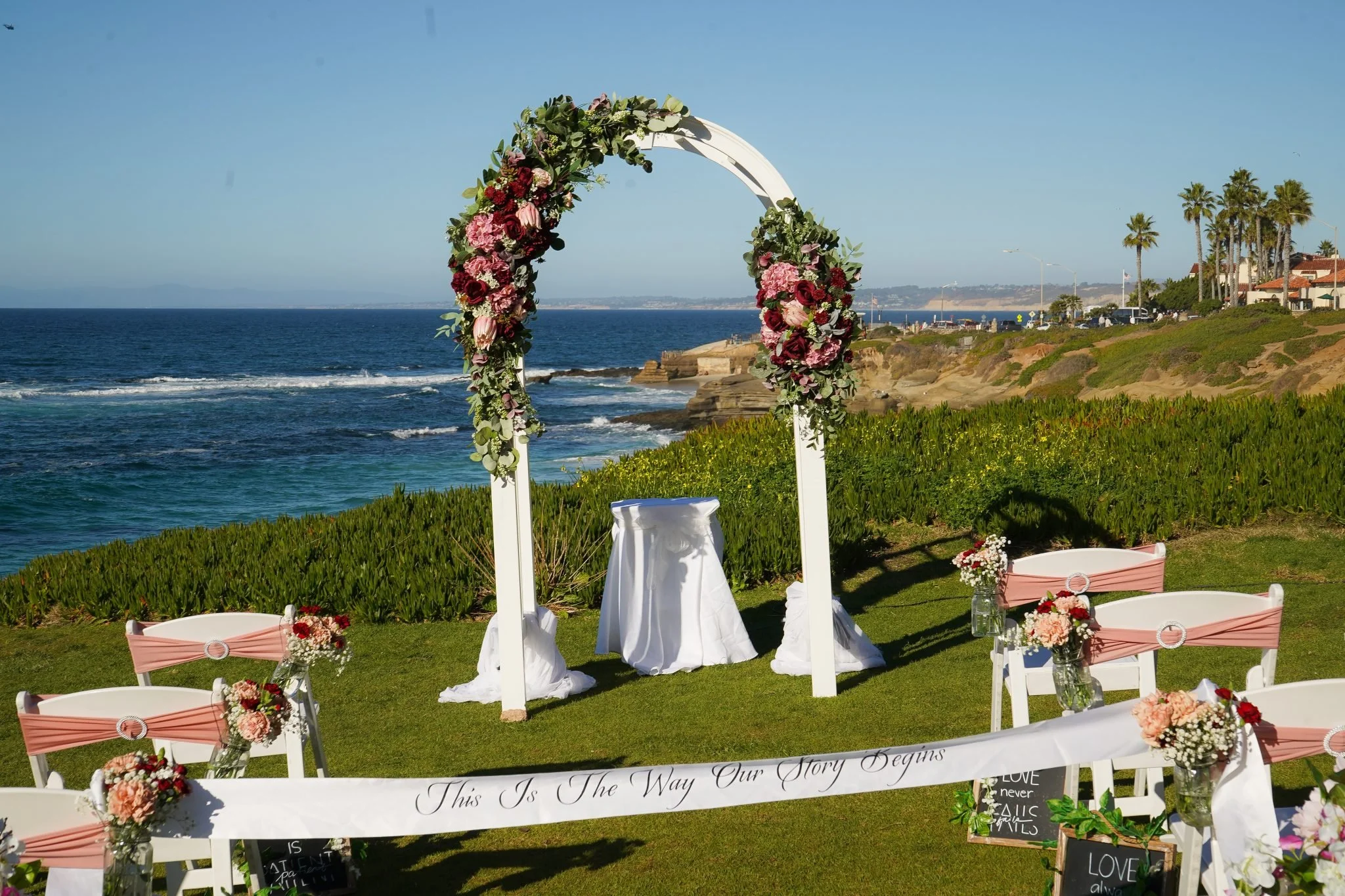 A wedding arch decorated with pink and red flowers and greenery, set up on a grassy area overlooking the ocean, with chairs and floral arrangements nearby.