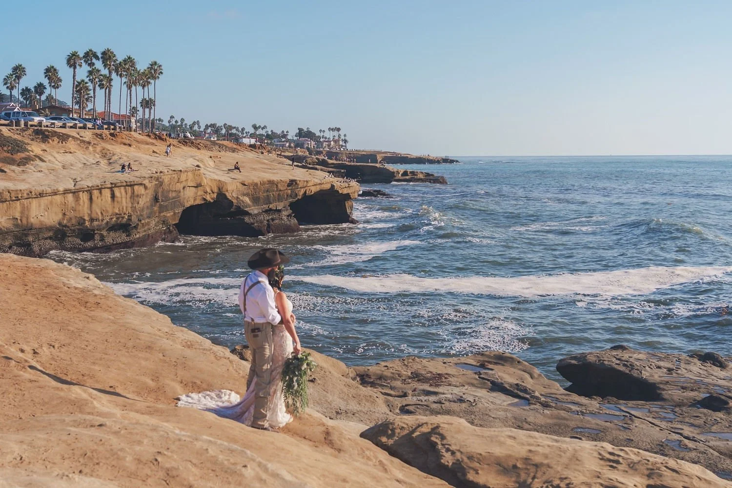 A couple getting married on a rocky beach with ocean waves, cliffs, palm trees, and parked cars in the background.