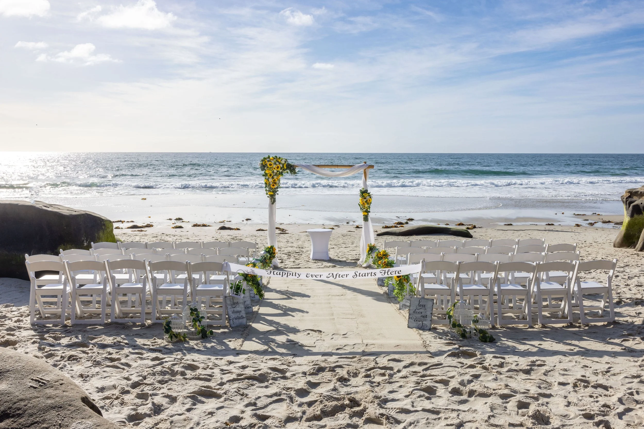 Beach wedding setup with white chairs facing an arch decorated with yellow flowers and white fabric, on sandy shore with ocean and cloudy sky in background.