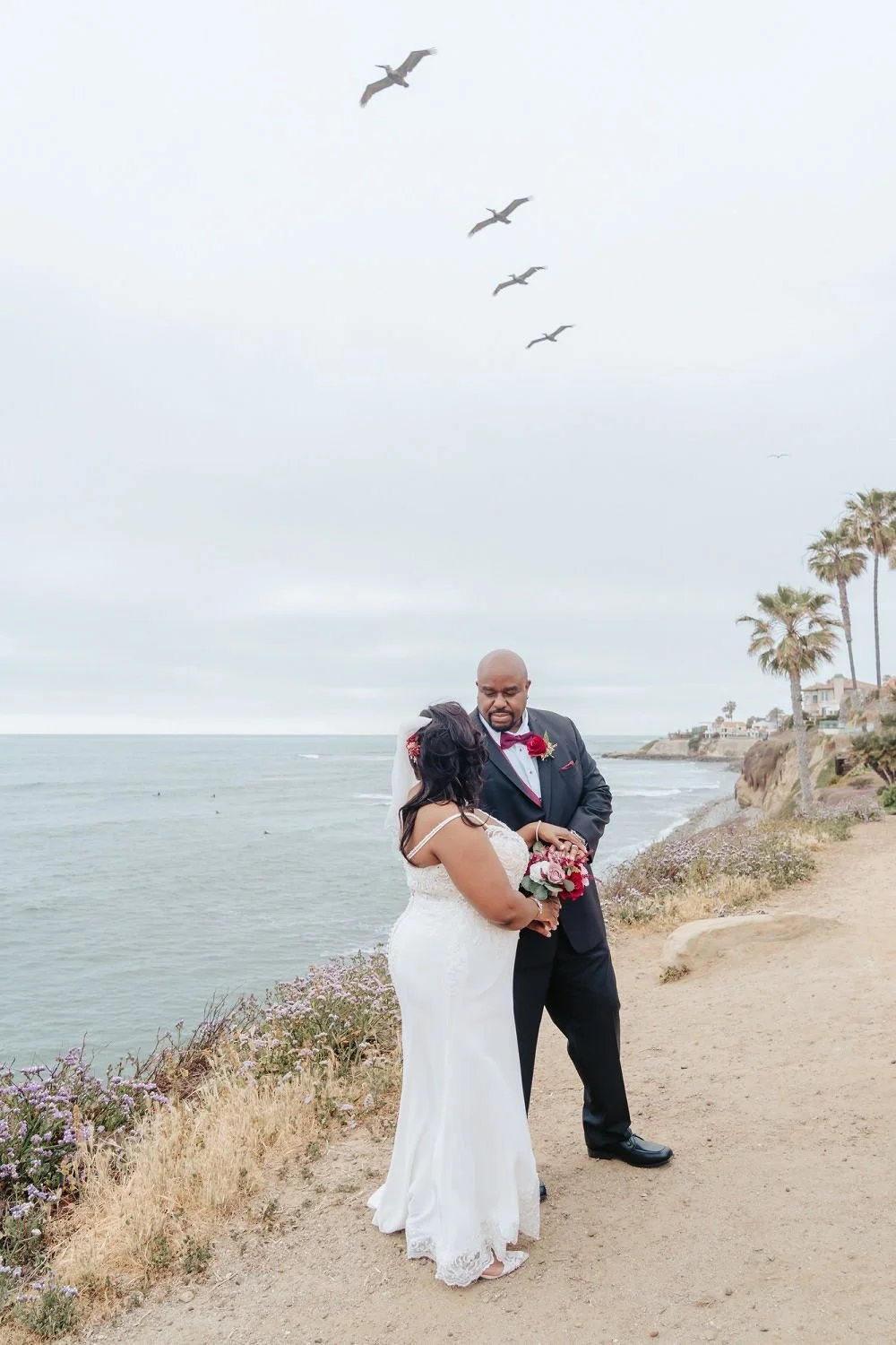 A couple dressed in wedding attire standing on a dirt path near the ocean, exchanging vows or rings, with seagulls flying overhead and palm trees in the background.