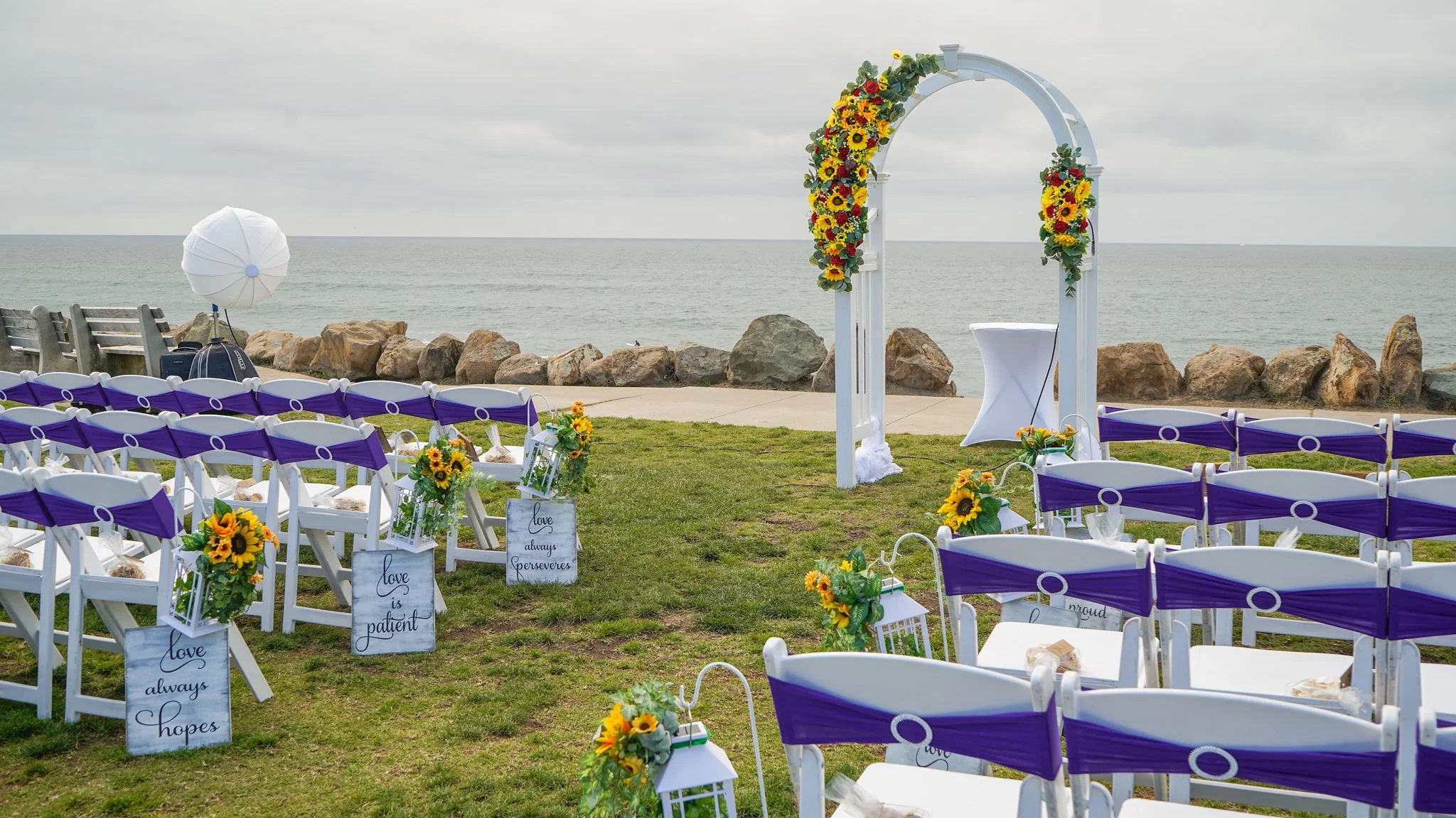Beachside wedding setup with white chairs decorated with sunflower arrangements, facing a white floral arch decorated with sunflowers, overlooking the ocean on a cloudy day.