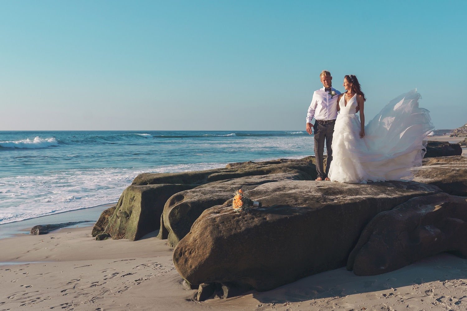 A newlywed couple stands on large rocks by the ocean, with the bride in a flowing white wedding dress and the groom in a white shirt and gray pants, overlooking the beach and waves.