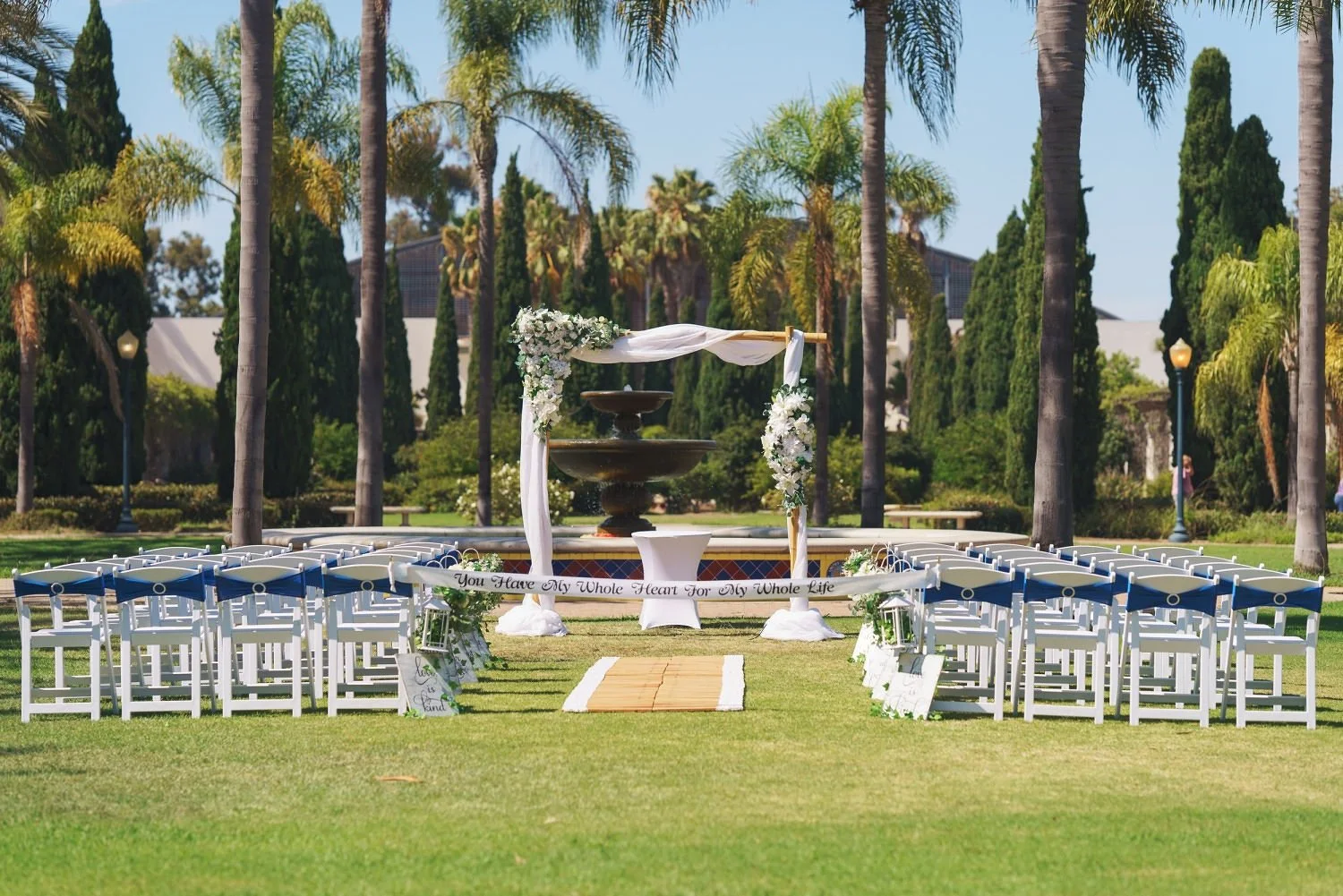 Outdoor wedding ceremony setup with white chairs, floral decorations, and an arch in front of a fountain, surrounded by palm trees and greenery.