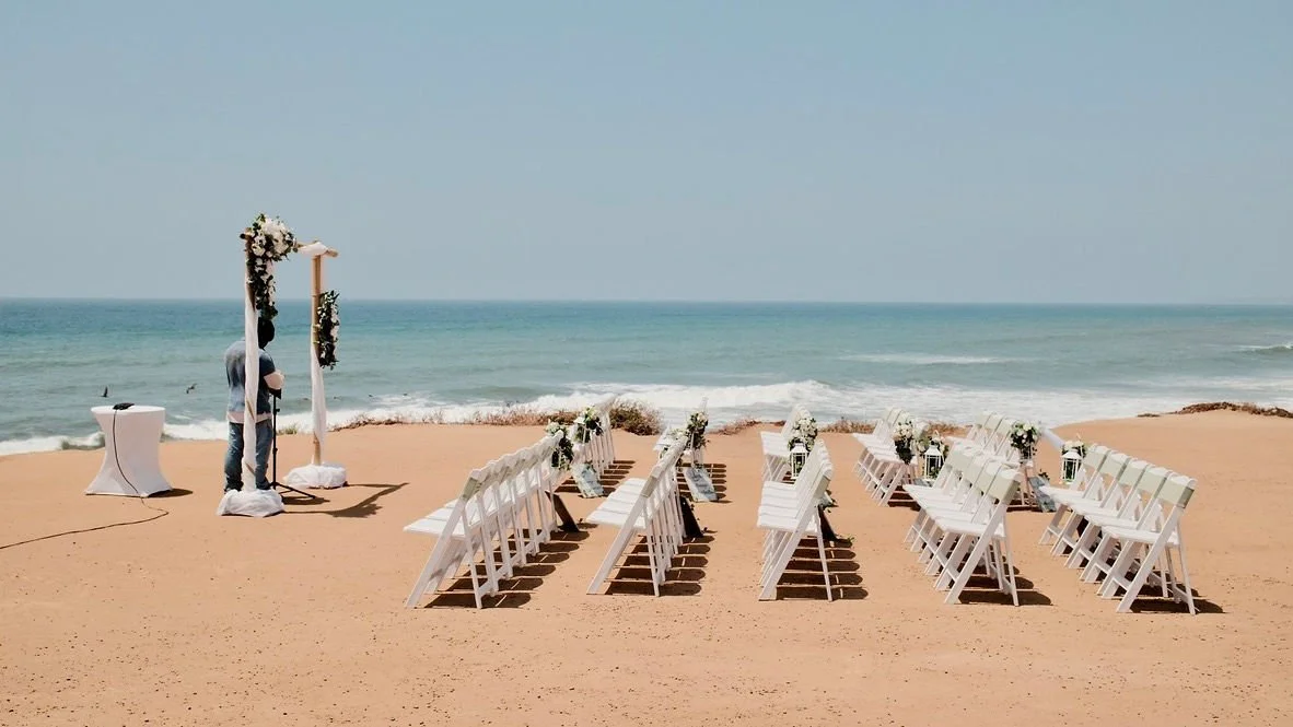 Empty chairs decorated for a beach wedding ceremony facing the ocean with a floral archway.
