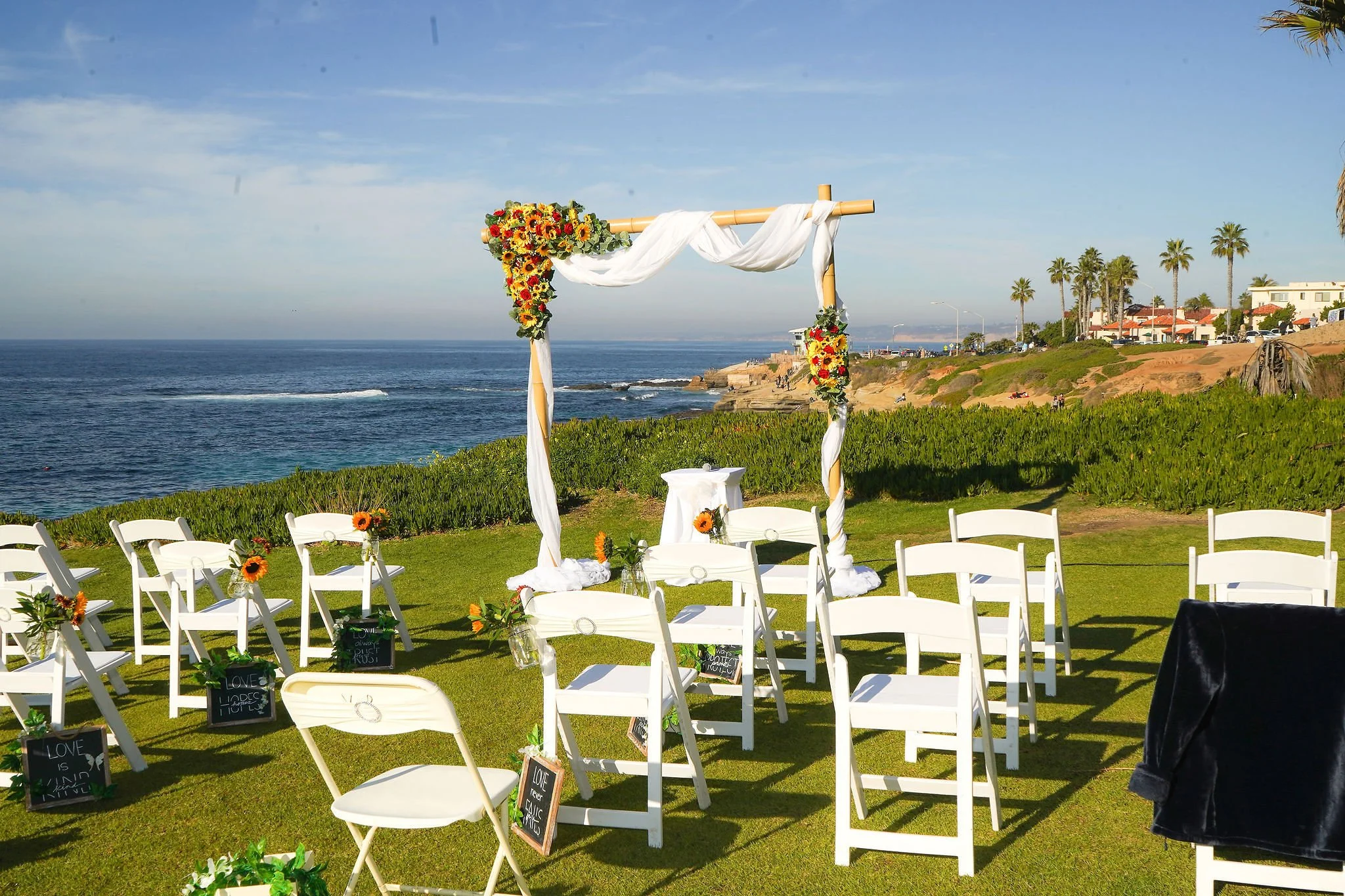 A decorated wedding ceremony setup on a grassy area overlooking the ocean. There are white chairs with small blackboards on the ground in front and a bamboo arch with white fabric and sunflower arrangements. The background features a blue sky, ocean 