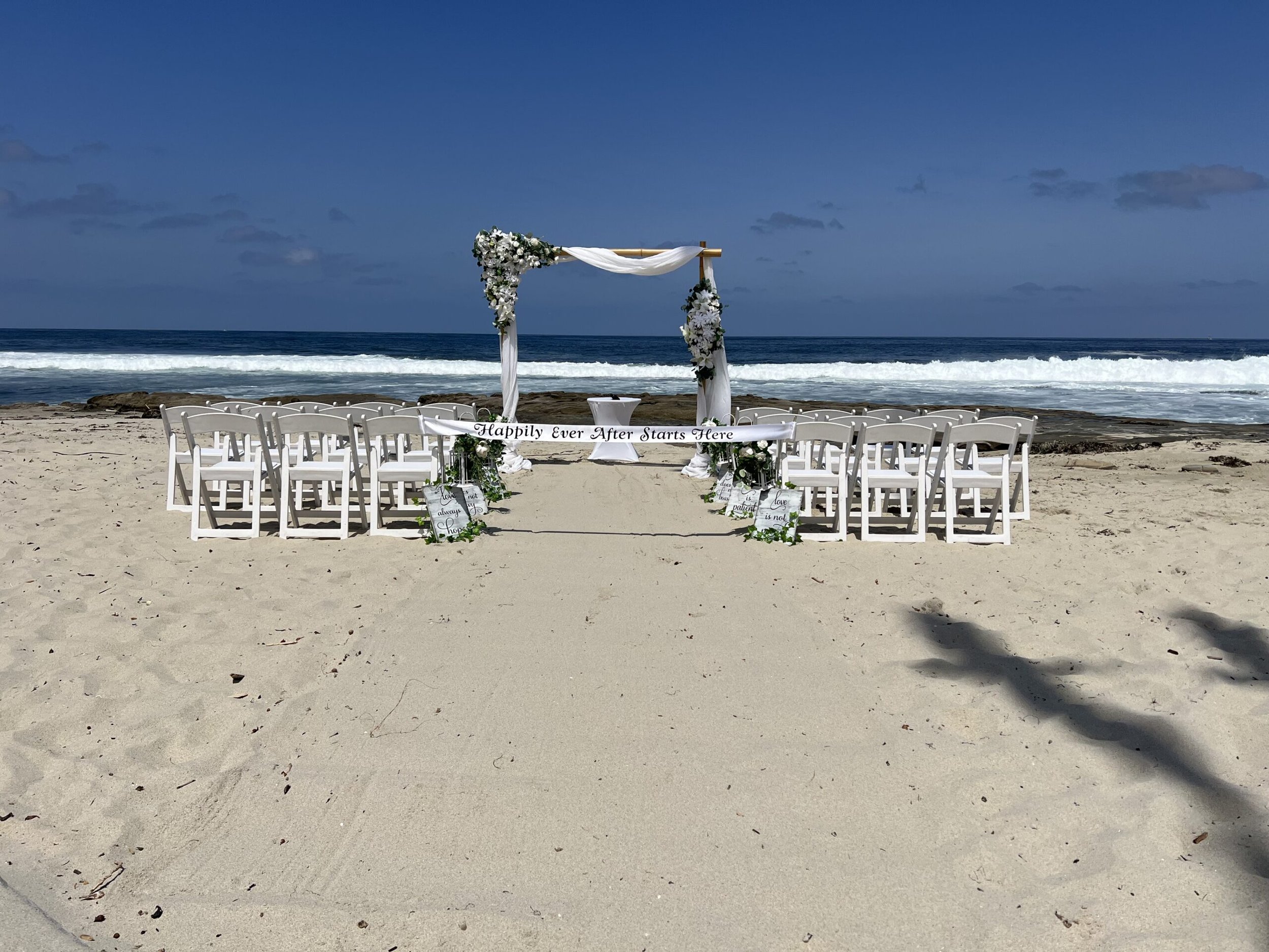 A wedding arch decorated with white flowers and white fabric, with white chairs arranged in rows on a sandy beach facing the ocean, under a partly cloudy sky with calm waves.