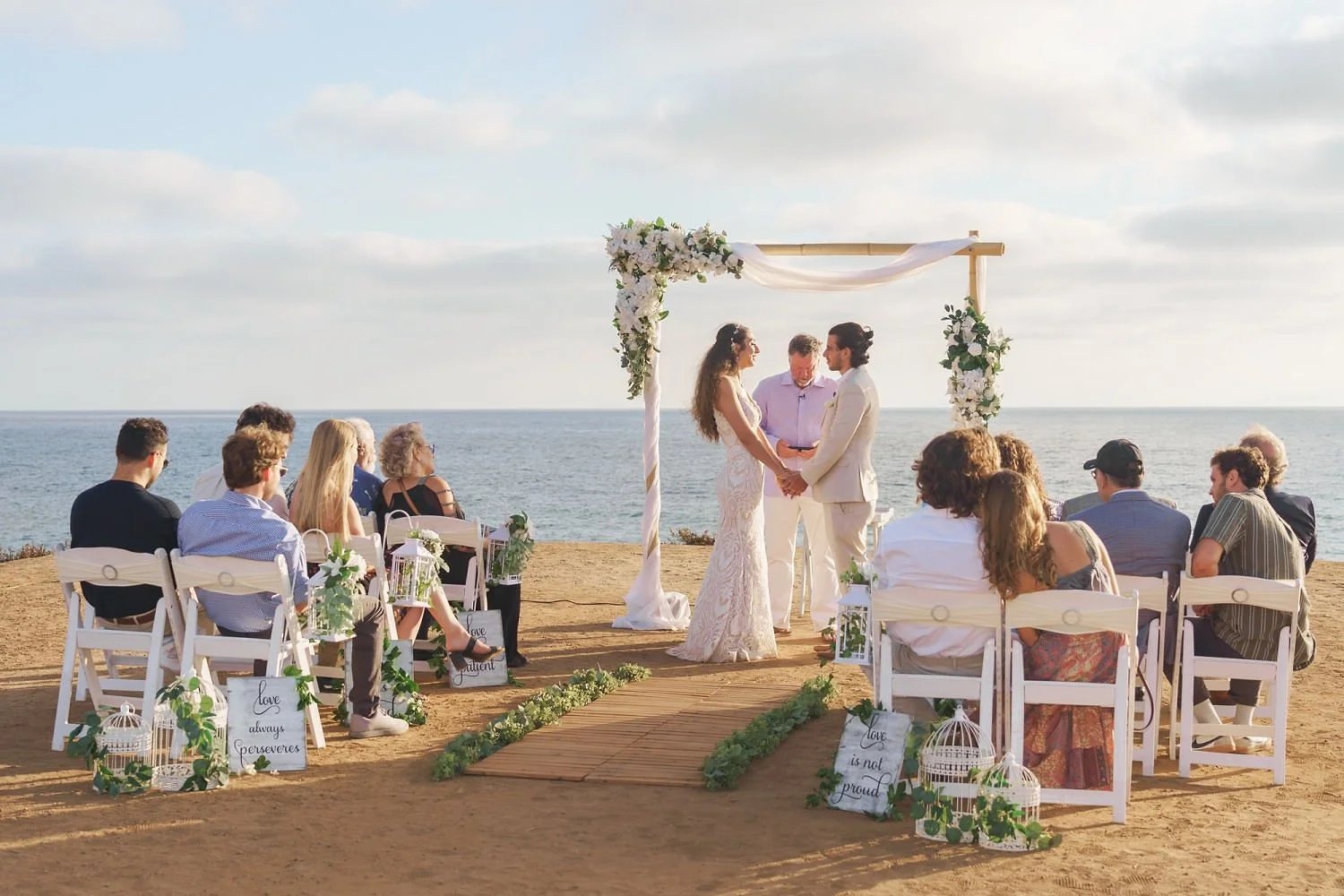 A wedding ceremony taking place on a beach with the ocean in the background, featuring a couple exchanging vows under a decorated arch, surrounded by seated guests and decorative signs and flowers.