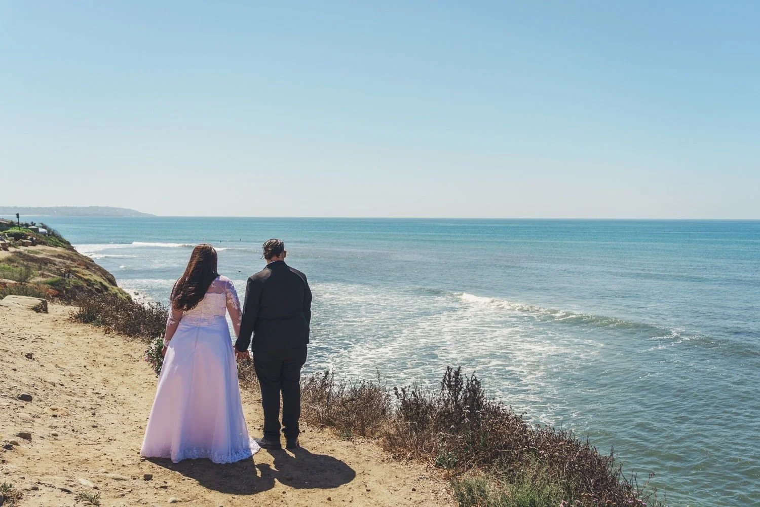 A bride and groom holding hands walking along a sandy coastal path near the ocean with clear blue skies.