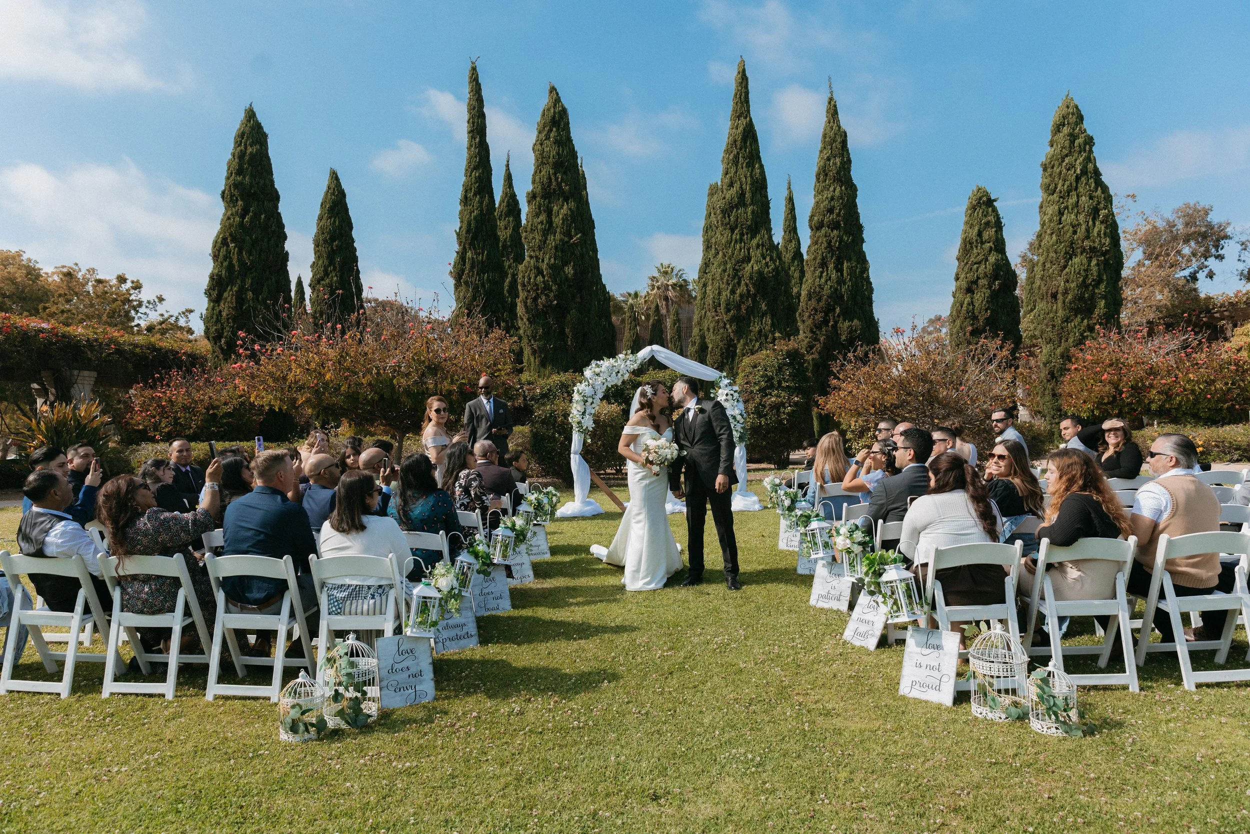 A wedding ceremony taking place outdoors in a garden with tall, narrow cypress trees in the background. The bride and groom are kissing under a floral arch, surrounded by guests seated in white chairs. Decor includes lanterns and signs with inspirati