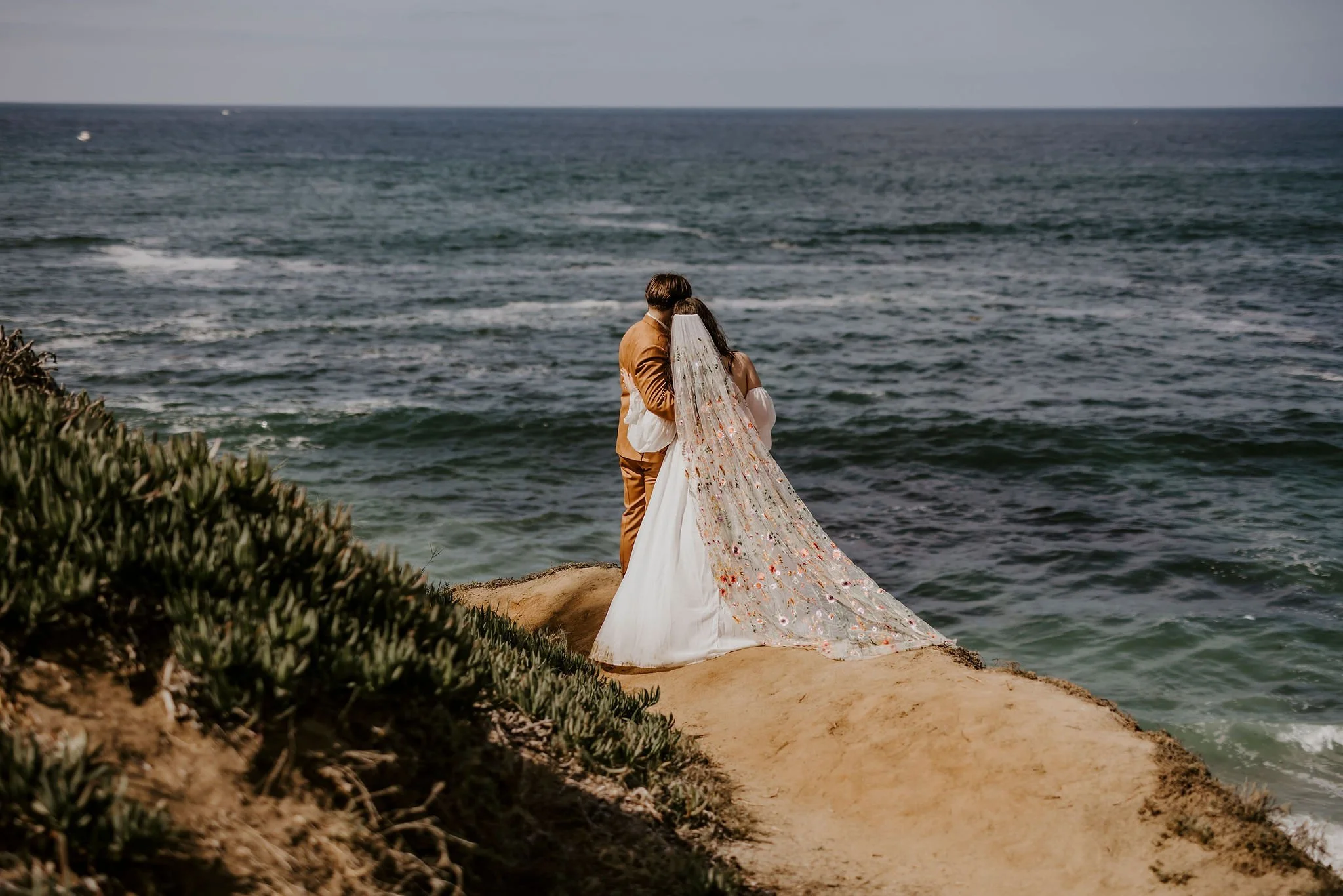 A couple dressed in wedding attire standing on a rocky beach, looking out at the ocean under a cloudy sky.
