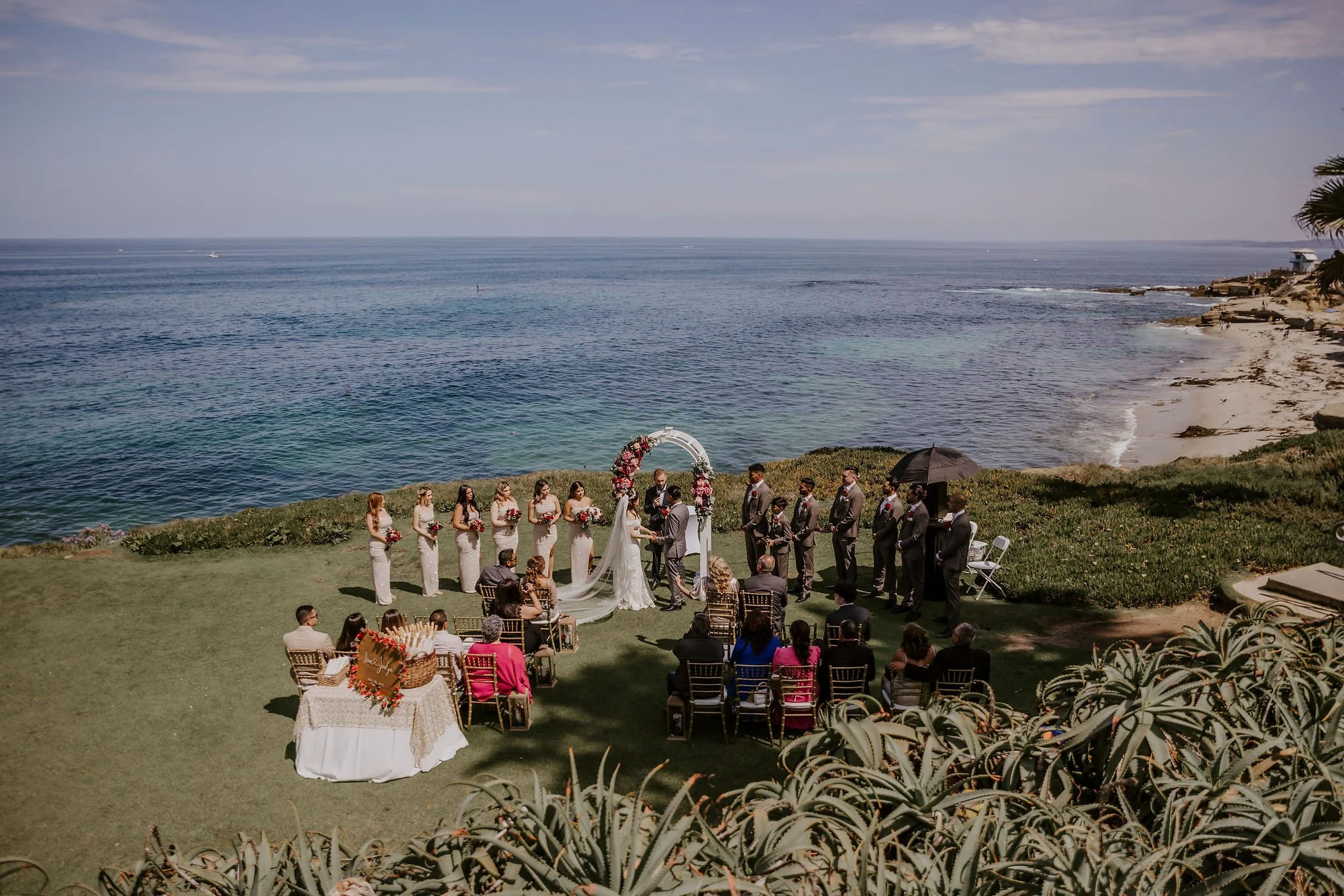 A wedding ceremony taking place outdoors on a grassy area near the ocean with guests seated, a bride and groom exchanging vows, and wedding party members standing nearby under a floral arch with the ocean and shoreline in the background.