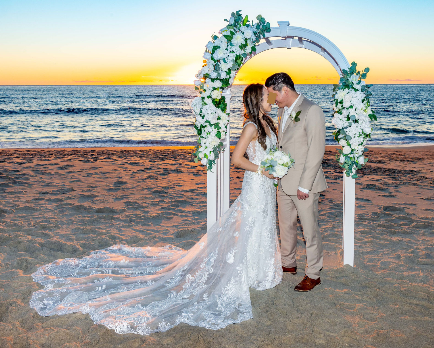 A bride and groom standing under a floral wedding arch on a beach during sunset.