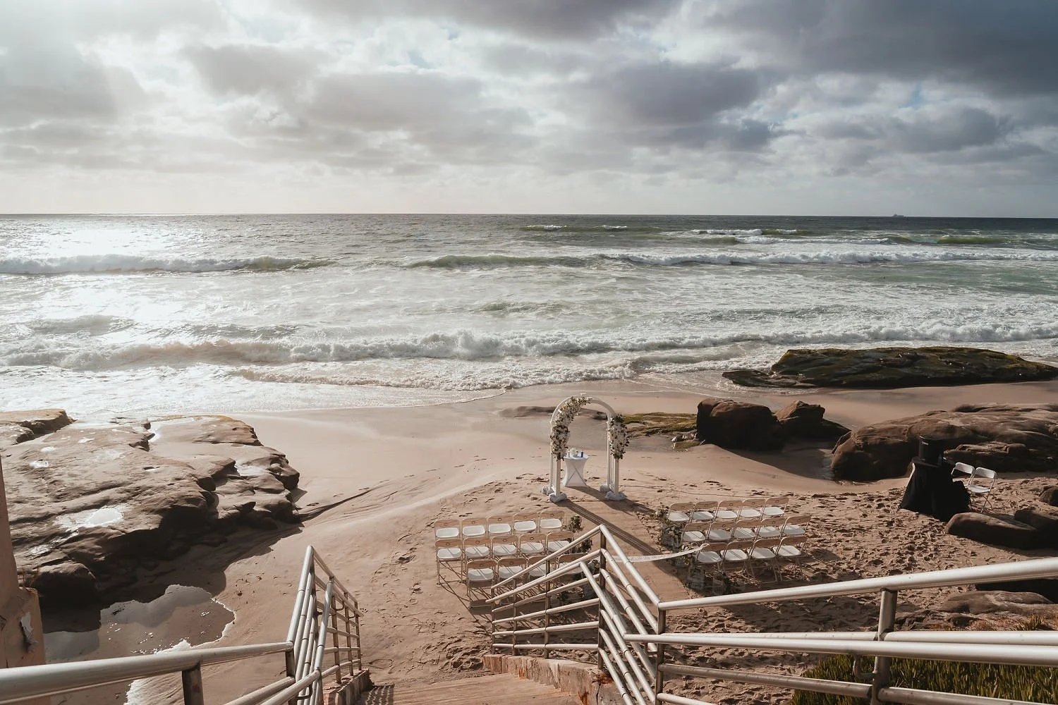 A beach setup for a wedding with white chairs arranged along the sand, an arch decorated with flowers, and the ocean in the background under cloudy skies.