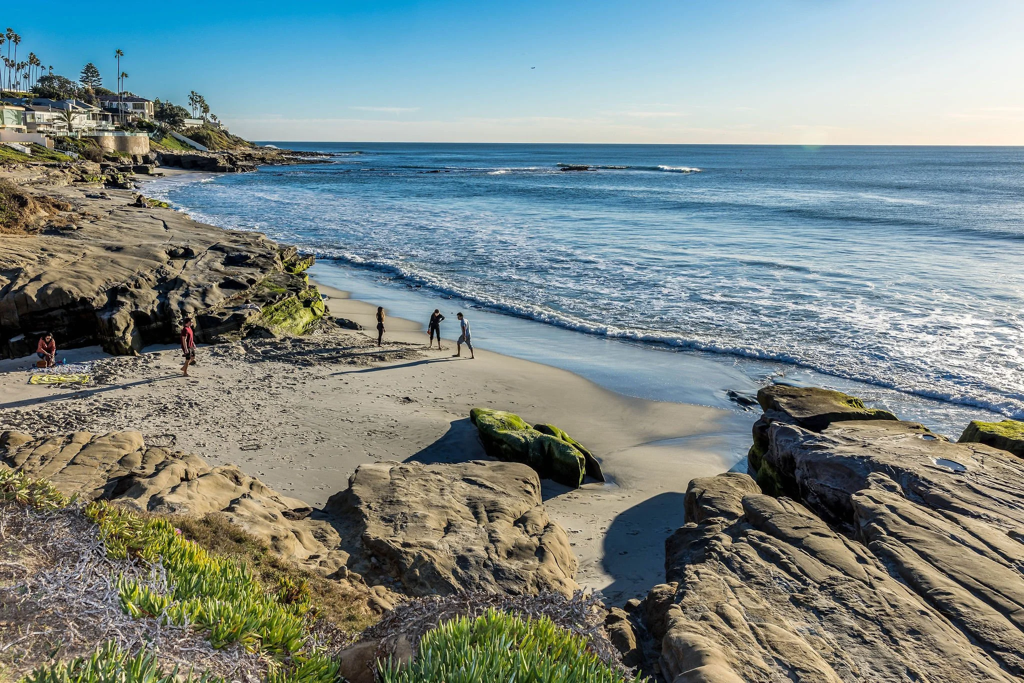 Beach with rocks, sand, and waves, houses on a hillside, and a few people walking or sitting near the shoreline in late afternoon sunlight.