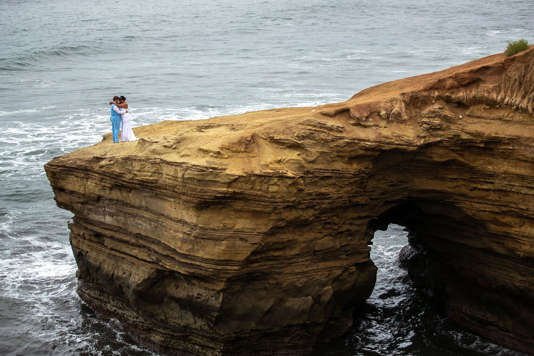 A couple embracing and kissing on a large rock formation by the ocean with waves crashing below.