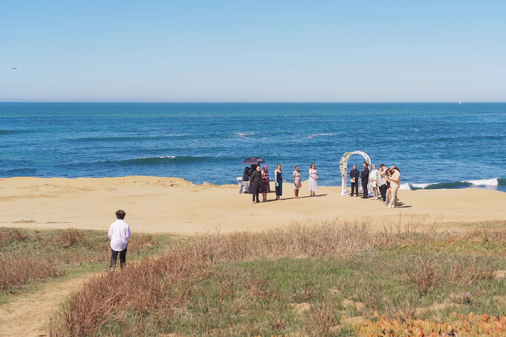 A beach wedding ceremony setup by the ocean with an arch decorated with flowers, guests standing nearby, and the sea and sky in the background.