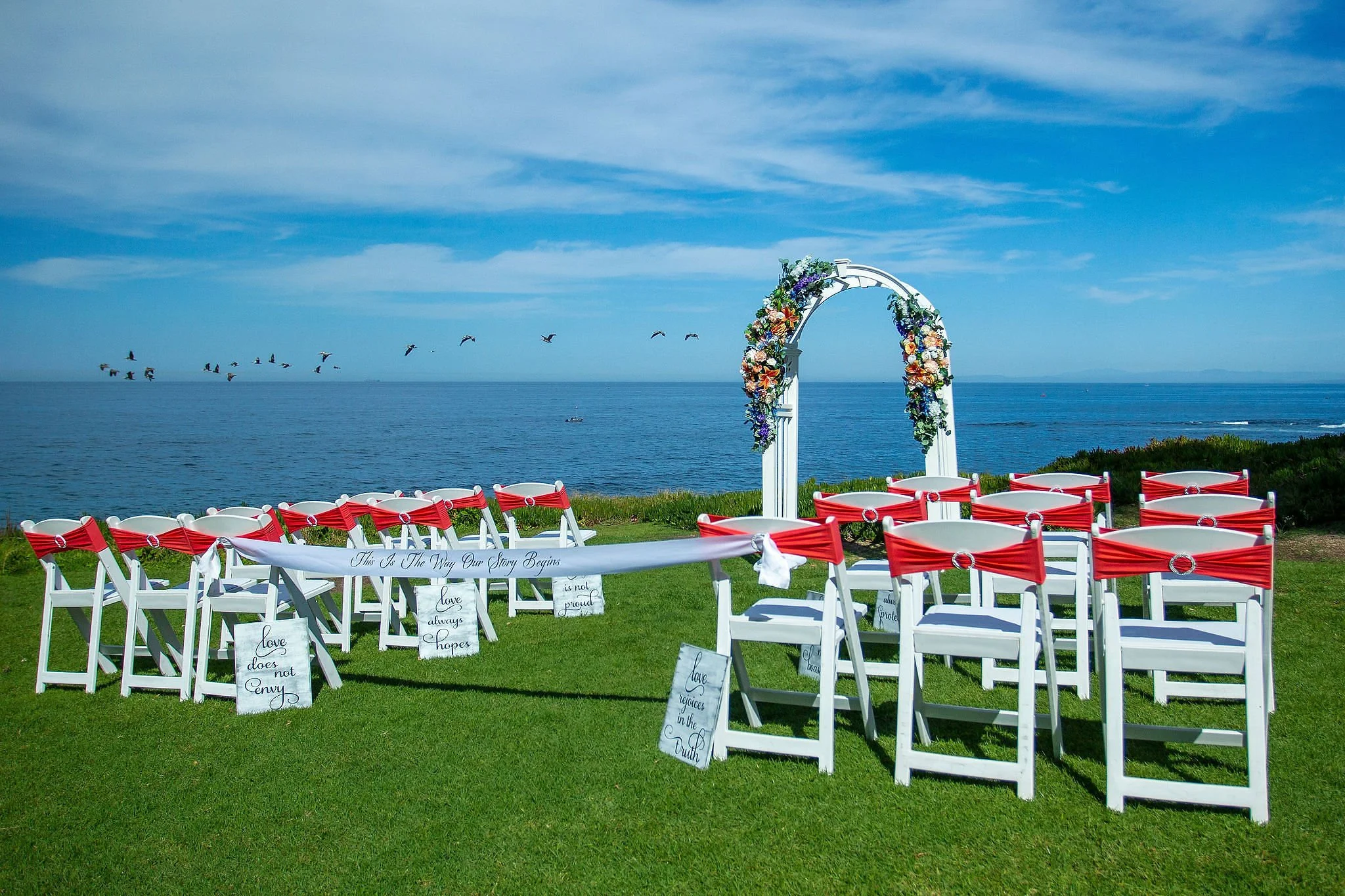 A seaside wedding setup with white chairs decorated with red ribbons, a floral arch, and a grassy area overlooking the ocean with a flock of birds flying in the sky.