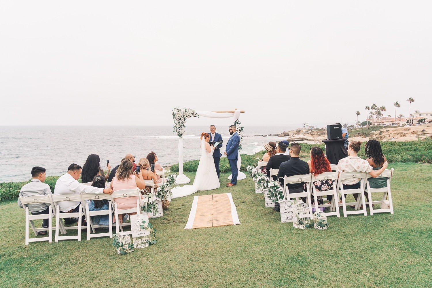 A beach wedding ceremony with a couple exchanging vows under a decorated arch, surrounded by seated guests, overlooking the ocean on a cloudy day.