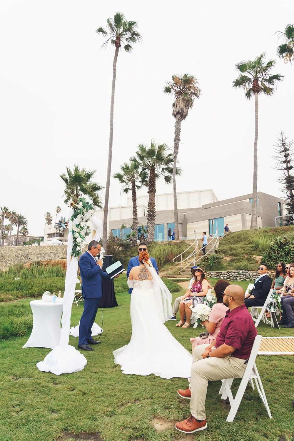 A wedding ceremony taking place outdoors under tall palm trees with guests seated on white chairs and a couple exchanging vows at an arch decorated with flowers.