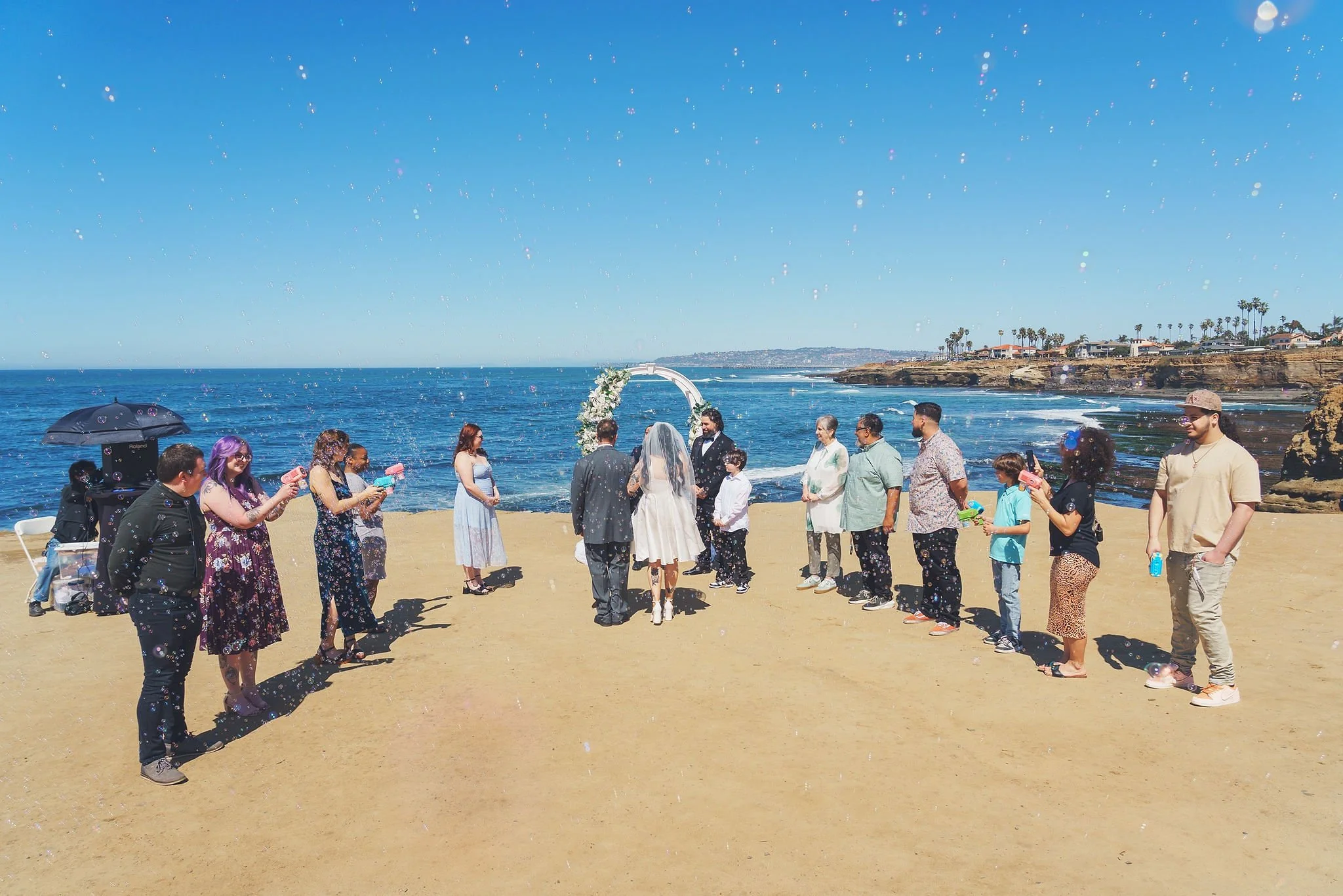 A group of people participating in a wedding ceremony on a beach, with an arch decorated with flowers, and the ocean and coastline in the background.