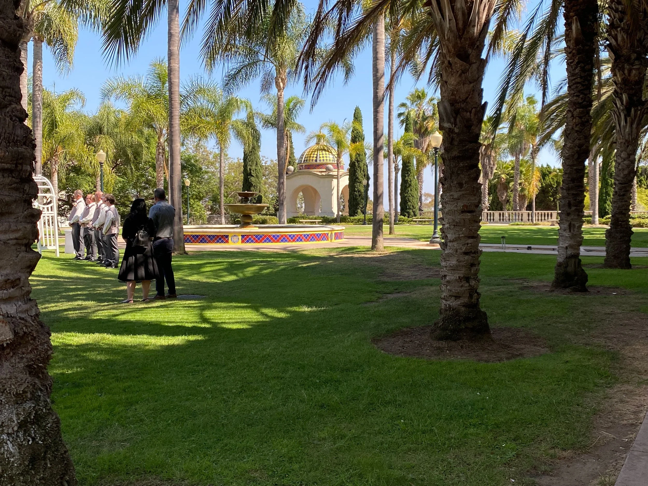 A group of people standing in a line outdoors among palm trees, with a decorative fountain and a small Pavilion in the background on a bright, sunny day.