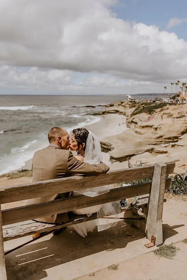 A bride and groom sitting on a wooden bench by the beach, sharing a kiss with ocean waves and cloudy skies in the background.