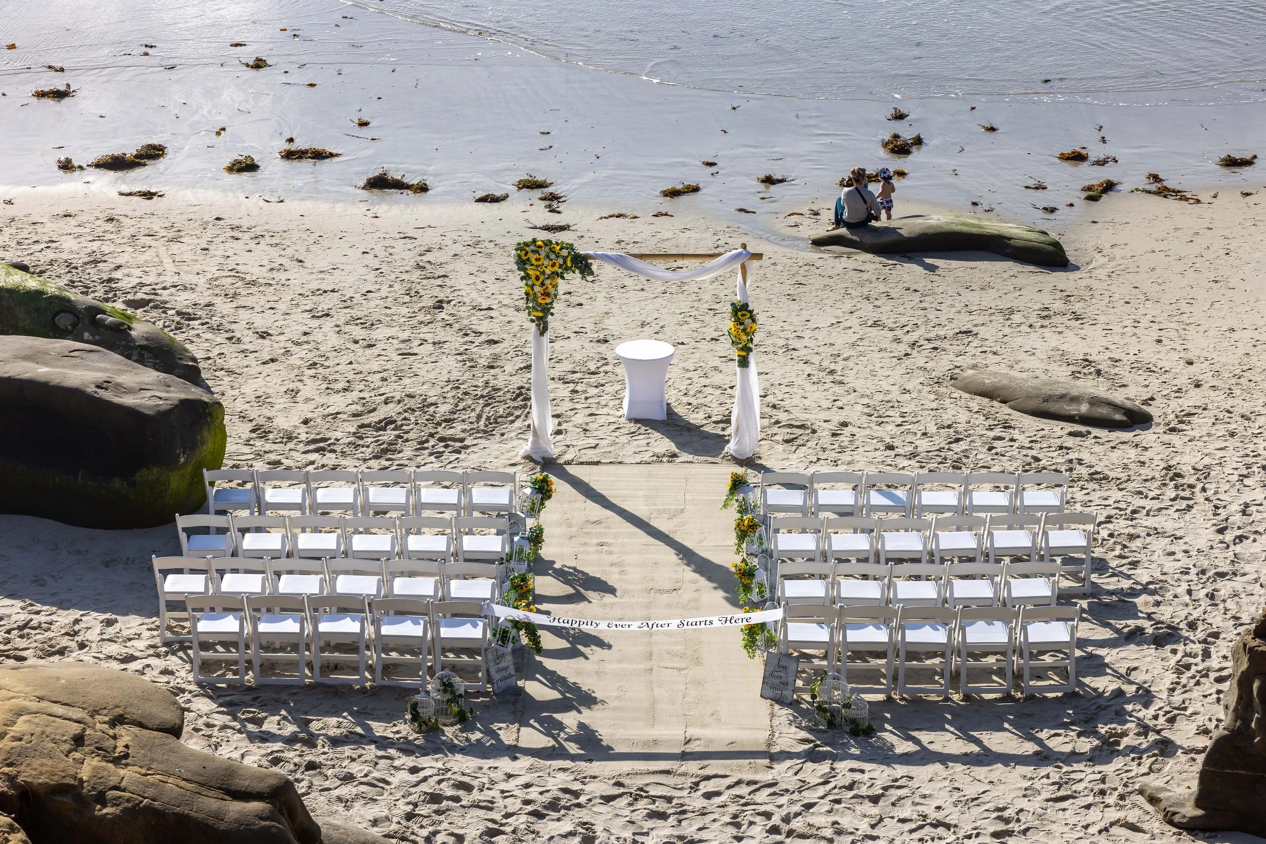 A beach wedding setup with white chairs arranged in rows facing the ocean, decorated with yellow flowers and white fabric, with a small arch draped in white fabric and yellow flowers, and a sign reading 'Happy ever After Starts Here.' The ocean is vi