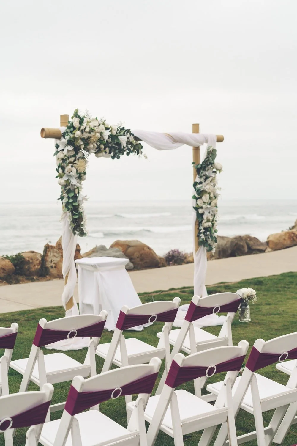 An outdoor wedding altar decorated with white flowers and draped fabric, set by the ocean with chairs arranged in front for guests.