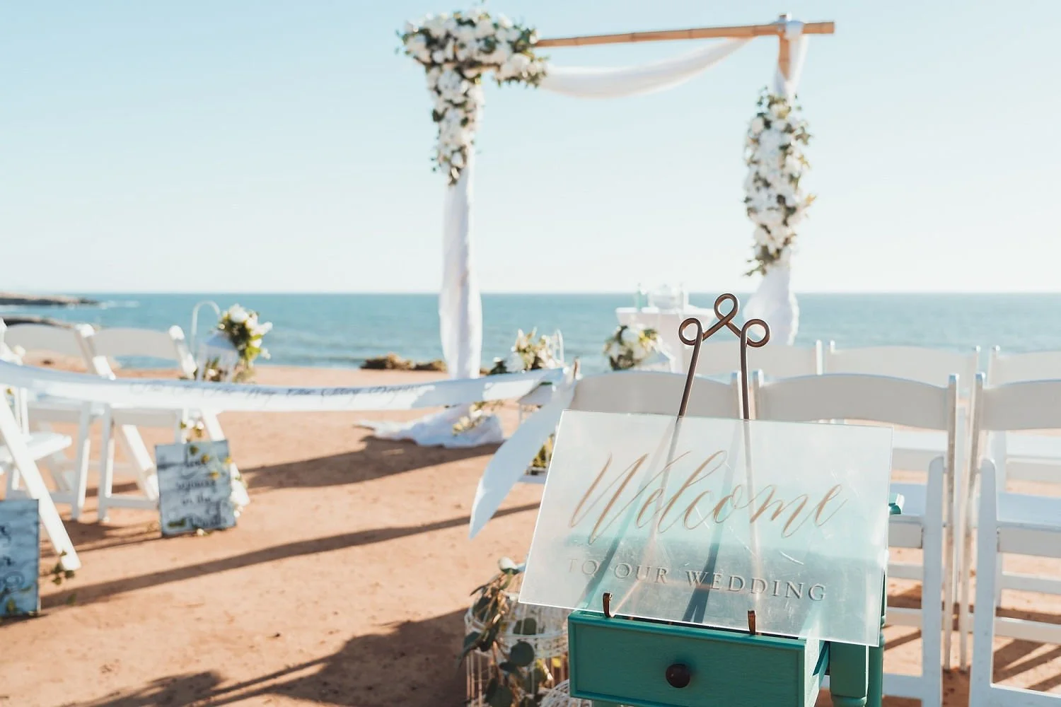 Wedding setup on a beach with an arch decorated with flowers, white chairs, a sign that says 'Welcome to our wedding', and the ocean in the background.
