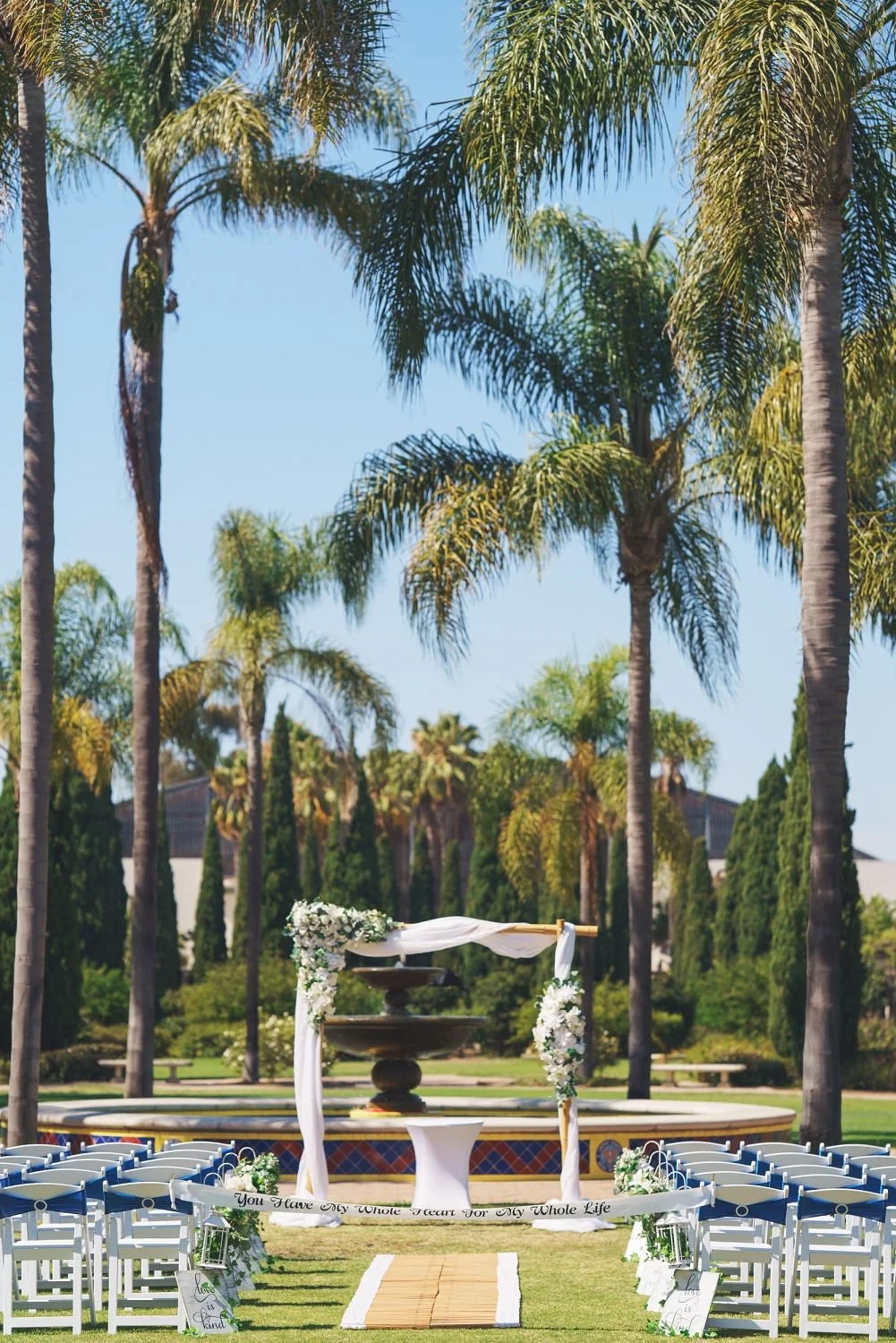 Outdoor wedding setup with chairs, floral decorations, and a ceremony arch under tall palm trees on a sunny day.