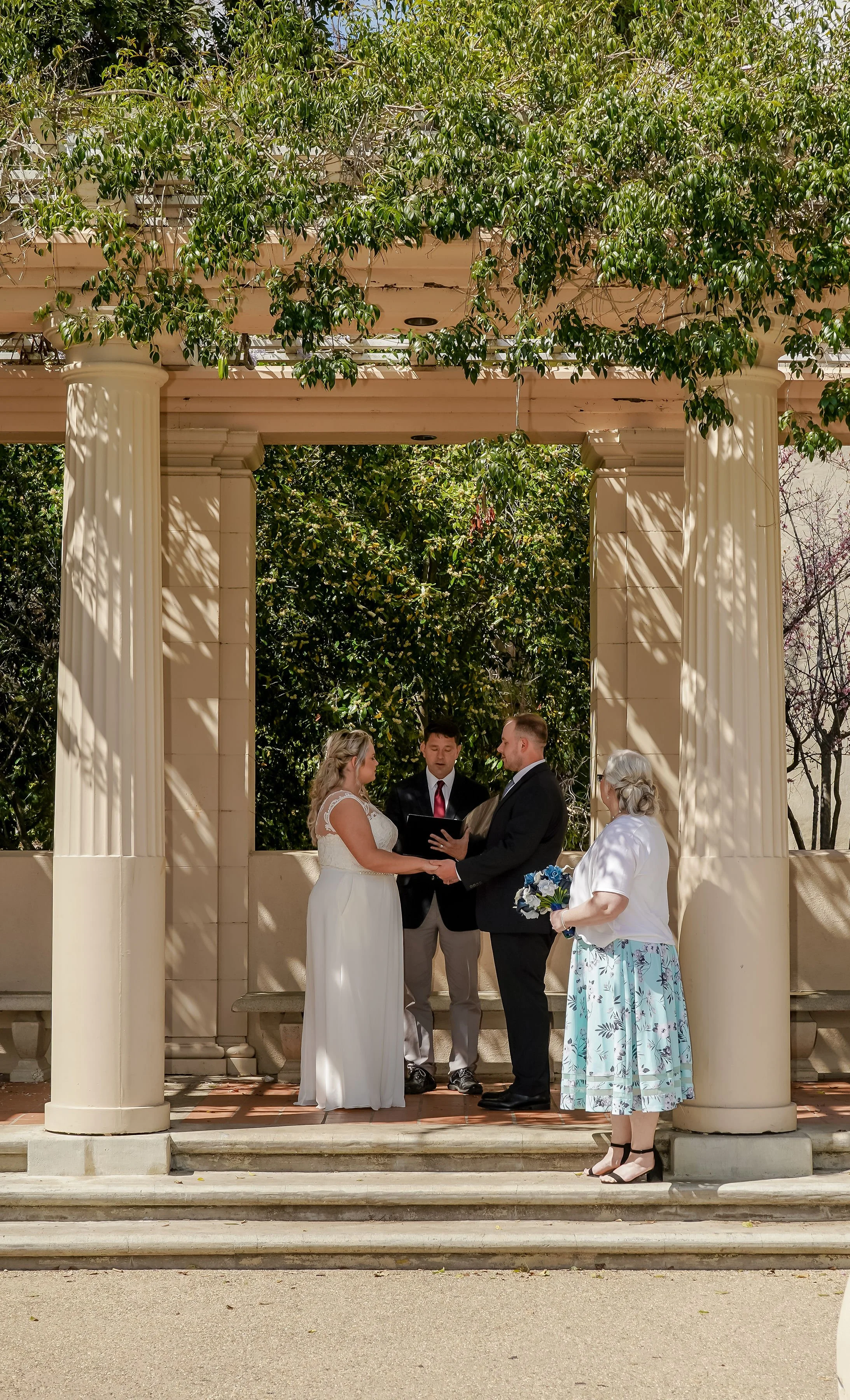 A wedding ceremony taking place outdoors under a classical-style pergola with columns and lush greenery. The bride and groom are holding hands and facing each other, with a officiant and a woman holding a bouquet nearby.