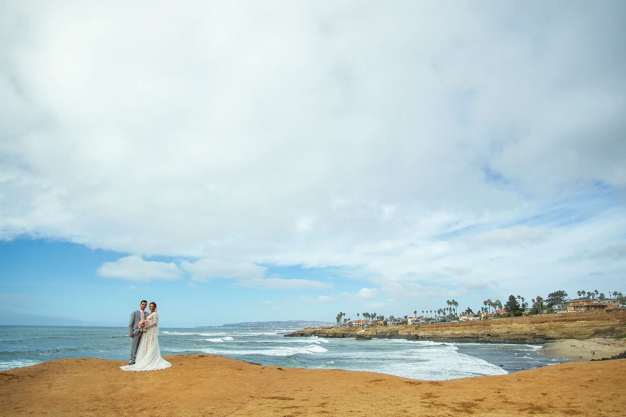 A couple dressed in wedding attire standing on a sandy beach near the ocean with a cloudy sky overhead and palm trees in the background.