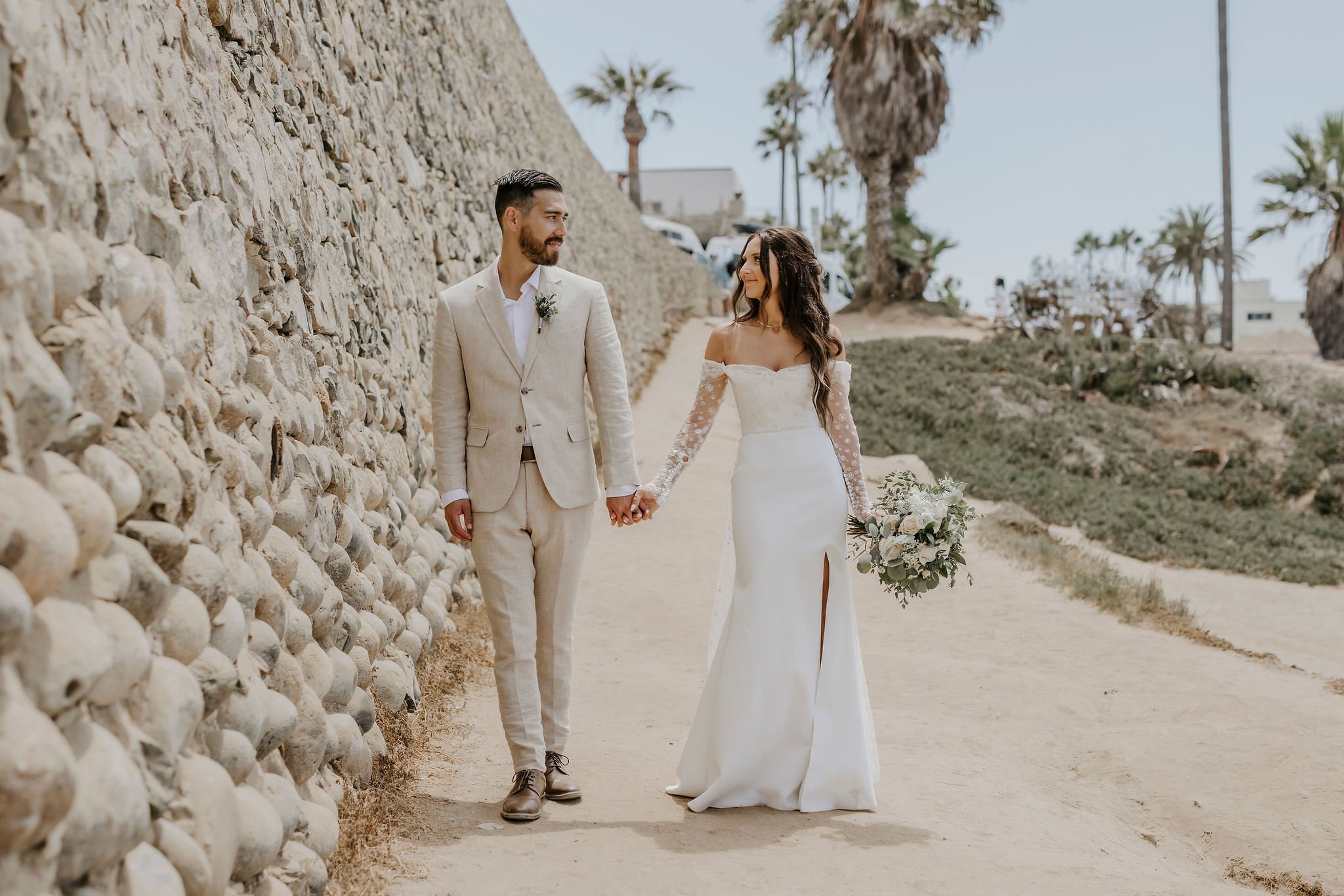A couple dressed in wedding attire holding hands and walking outdoors by a stone wall, with palm trees and clear sky in the background.