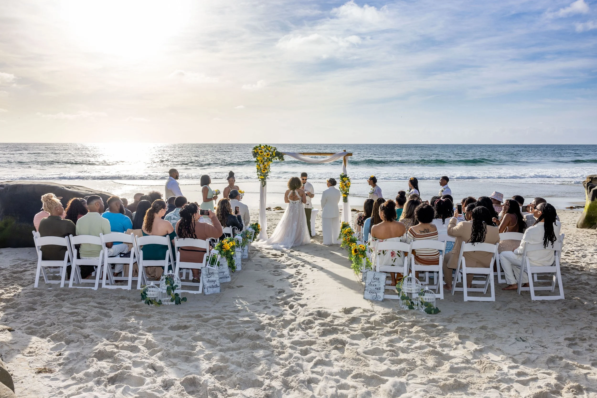 A beach wedding ceremony with a large gathering of guests seated on white chairs, facing an altar decorated with flowers and fabric, overlooking the ocean during sunset.