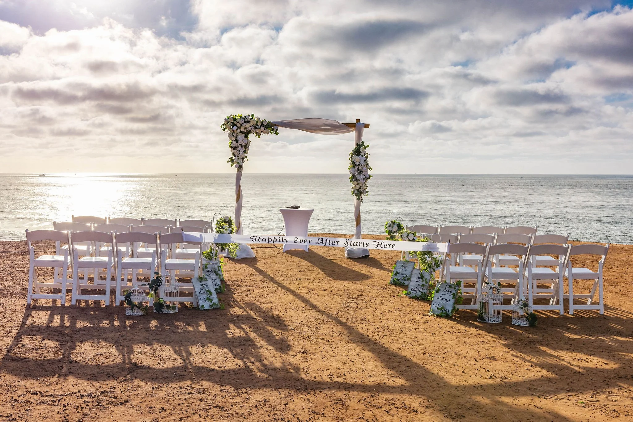Beachside wedding setup with white chairs, floral decorations, an arch draped with fabric and flowers, with the ocean and cloudy sky in the background.
