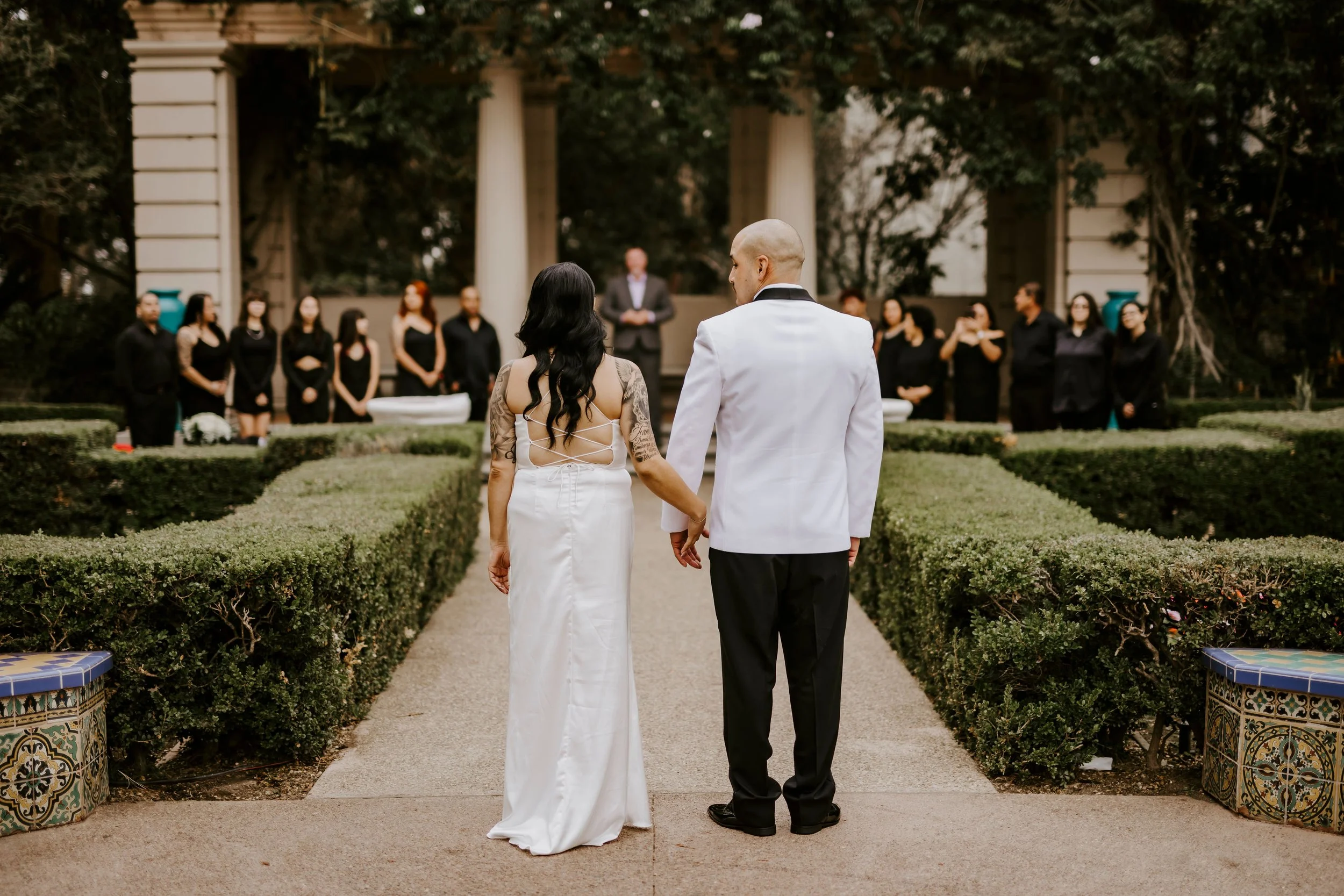 A couple getting married outdoors, holding hands, with a wedding officiant and wedding party in the background.