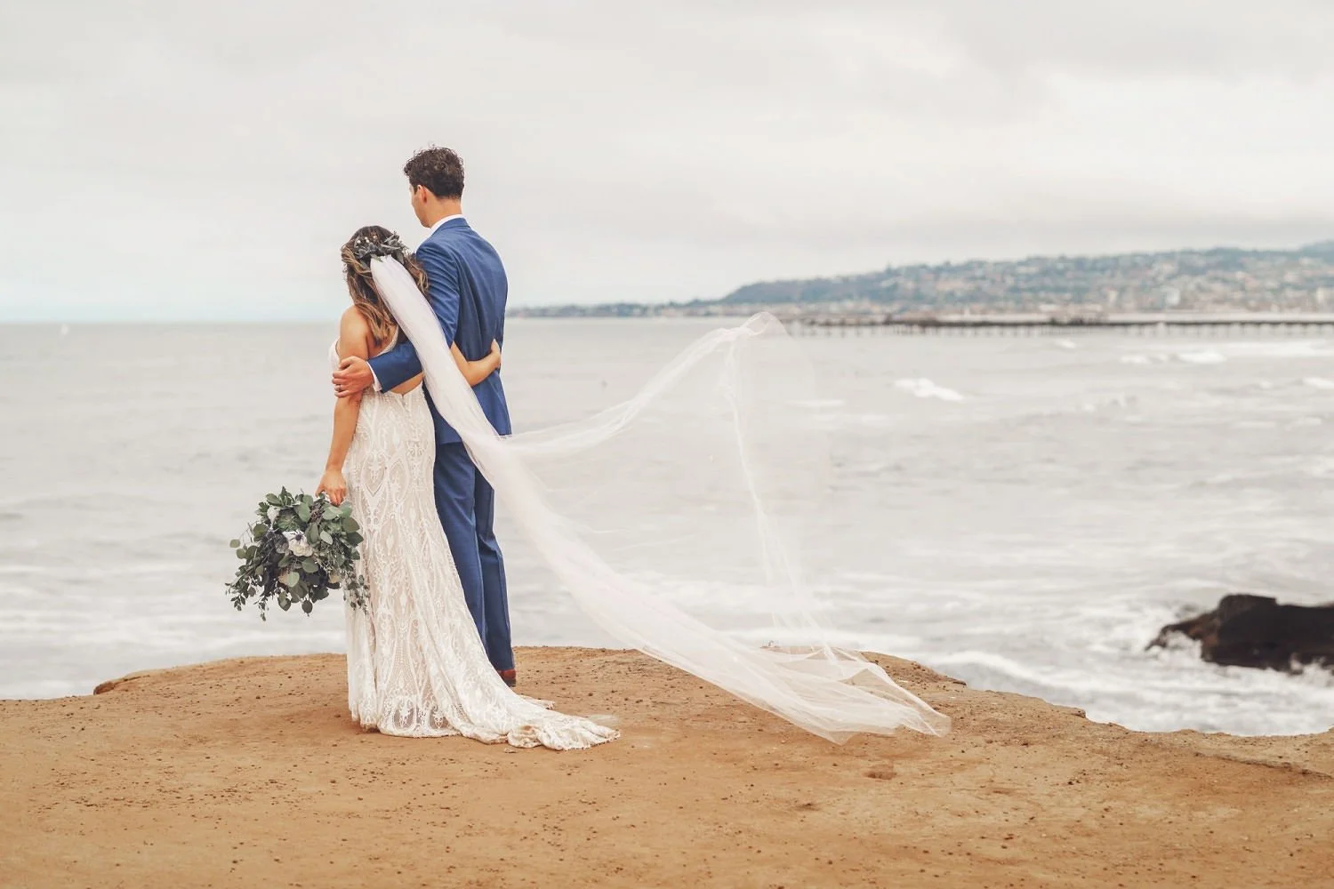 Bride and groom embracing on the shoreline after their intimate San Diego beach wedding ceremony, with ocean waves, coastal cliffs, and the bride’s long veil flowing in the breeze.