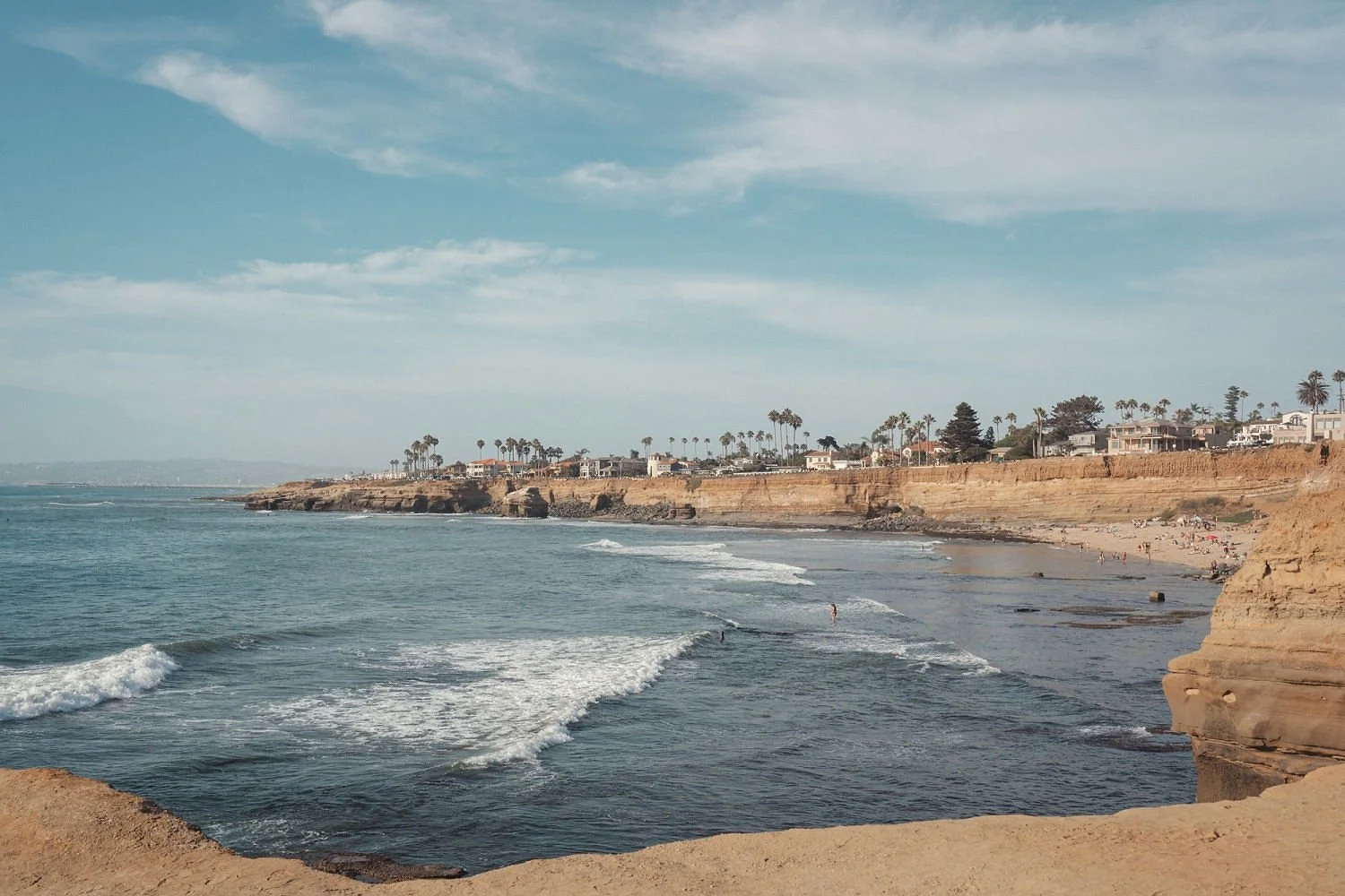 A scenic view of a beach with ocean waves, sandy cliffs, and houses with palm trees in the background, under a partly cloudy sky.