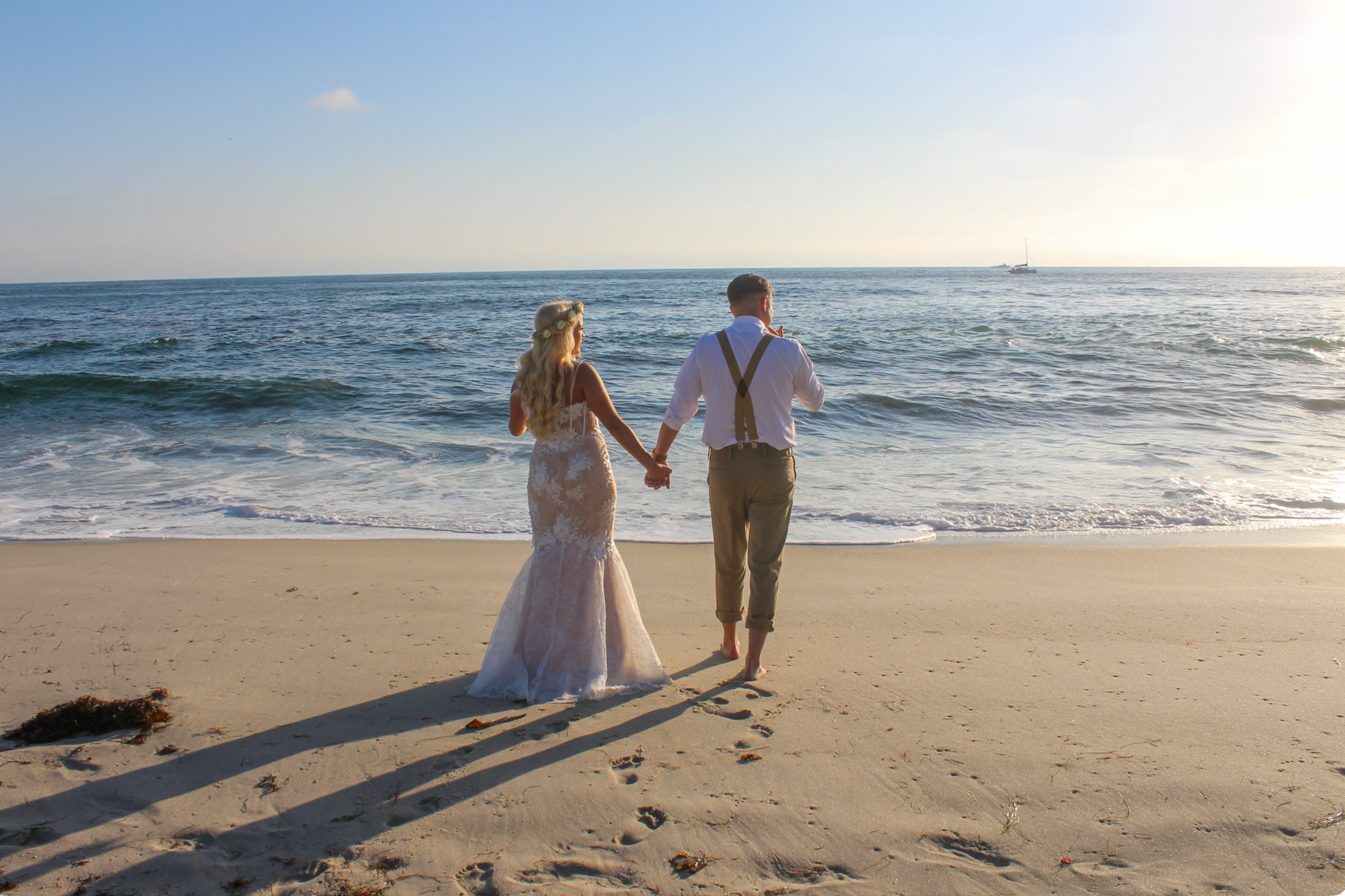 A bride and groom holding hands and walking on the beach near the ocean during sunset.