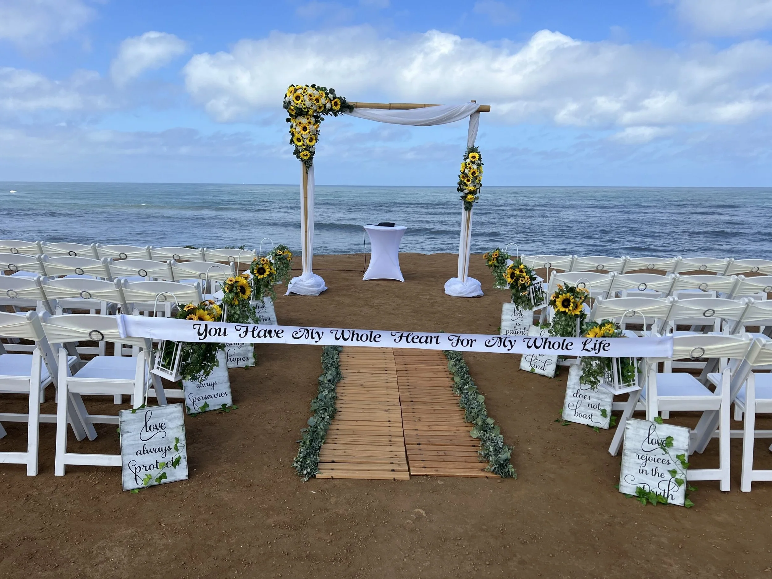 A wedding setup on a beach with white chairs arranged on either side of a wooden aisle. The aisle leads to an arch decorated with yellow sunflowers and white fabric, overlooking the ocean. Several signs with heartfelt messages about love and hope are placed along the aisle. The sky is partly cloudy.