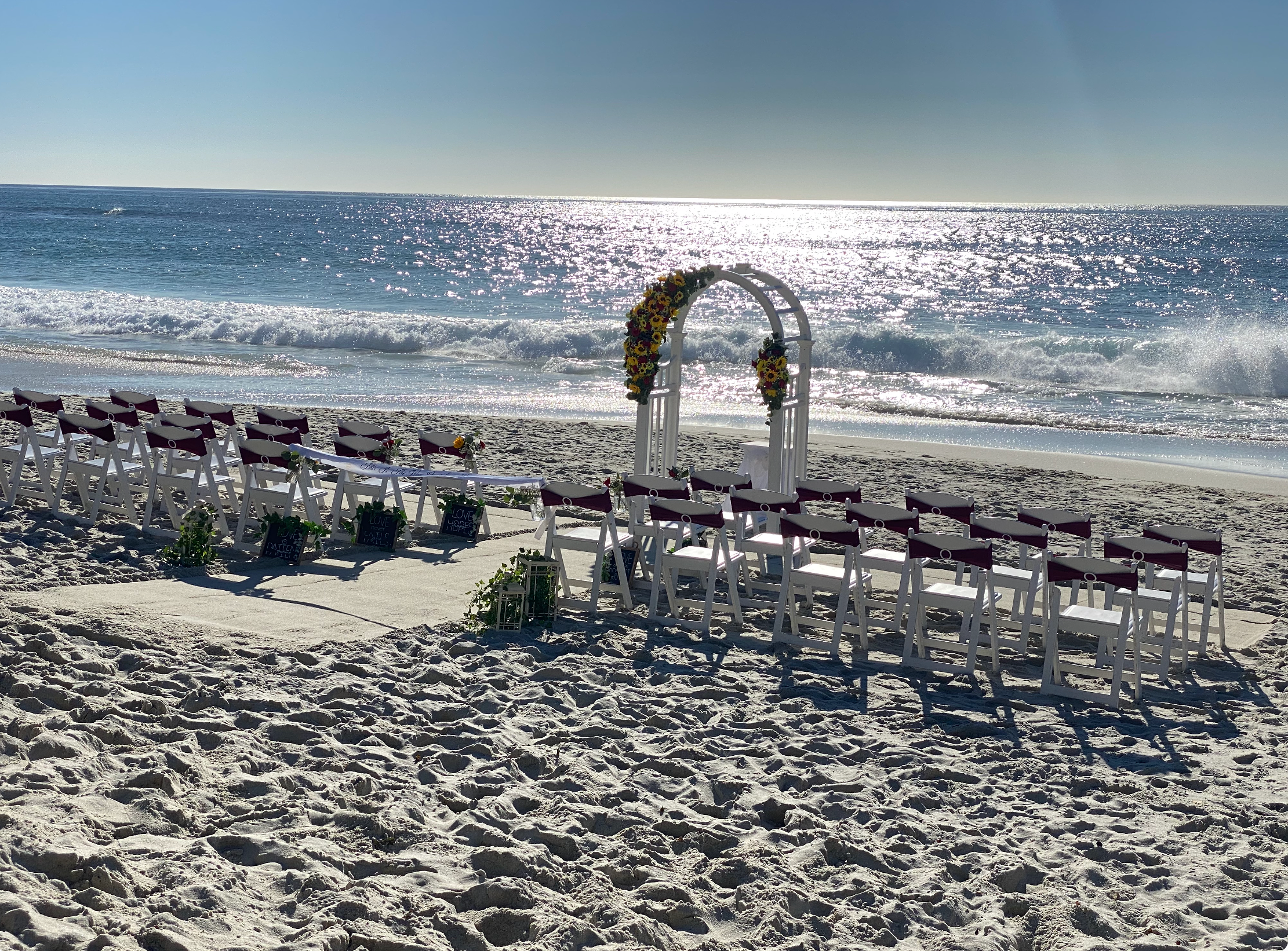 A wedding setup on a sandy beach with rows of chairs, a floral arch, and an ocean view in the background.