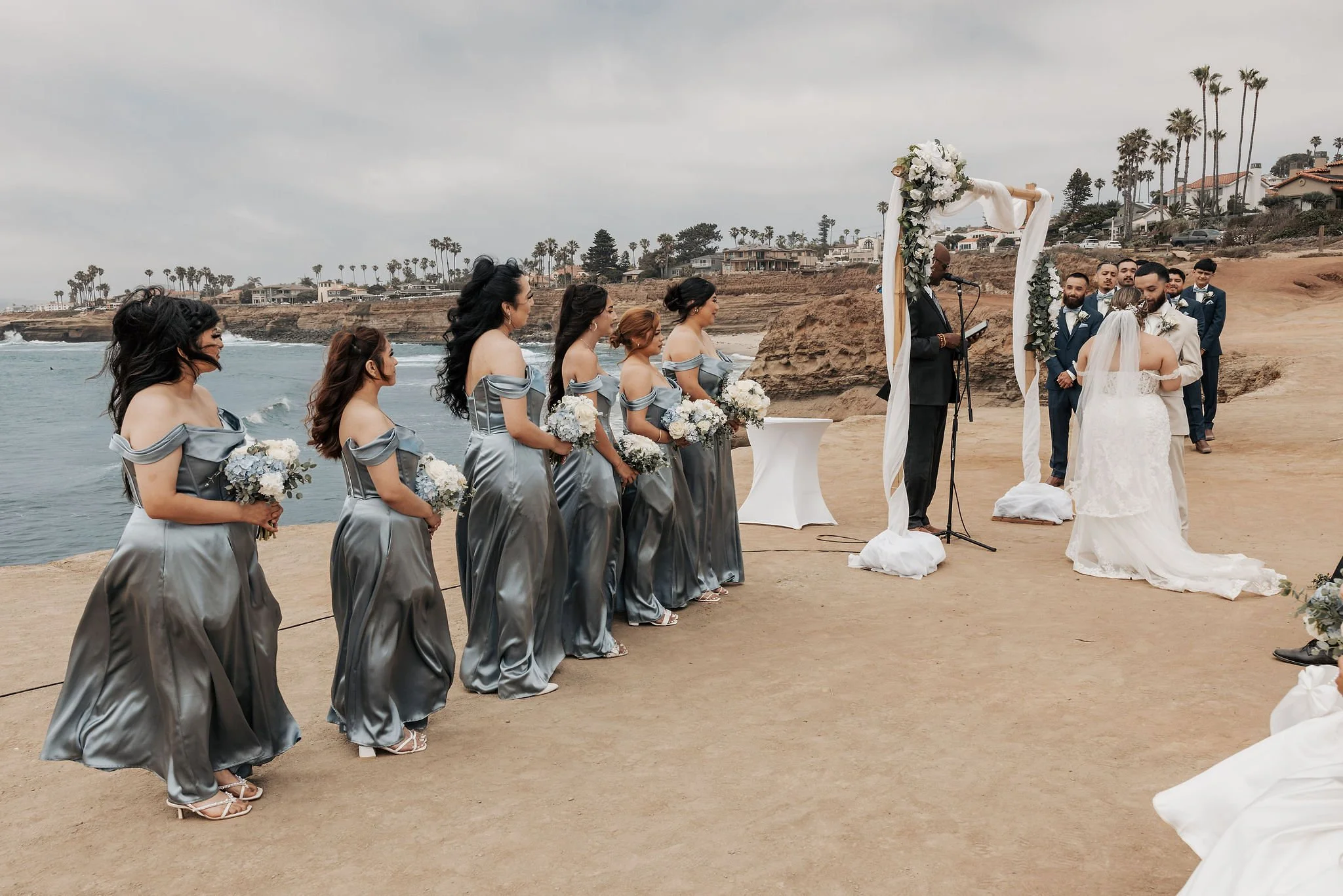 A beach wedding ceremony with bridesmaids in matching silver dresses holding bouquets, a bride and groom embracing at the altar, and an officiant speaking. The setting features rocky cliffs, palm trees, and houses in the background under a cloudy sky