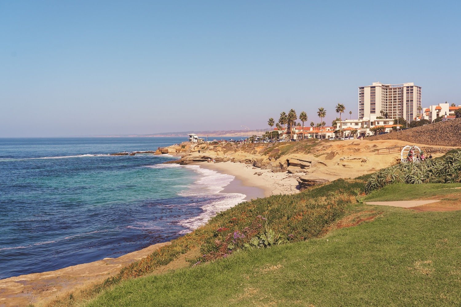 Coastal scene with sandy beach, ocean waves, cliffs, palm trees, and buildings in the background.