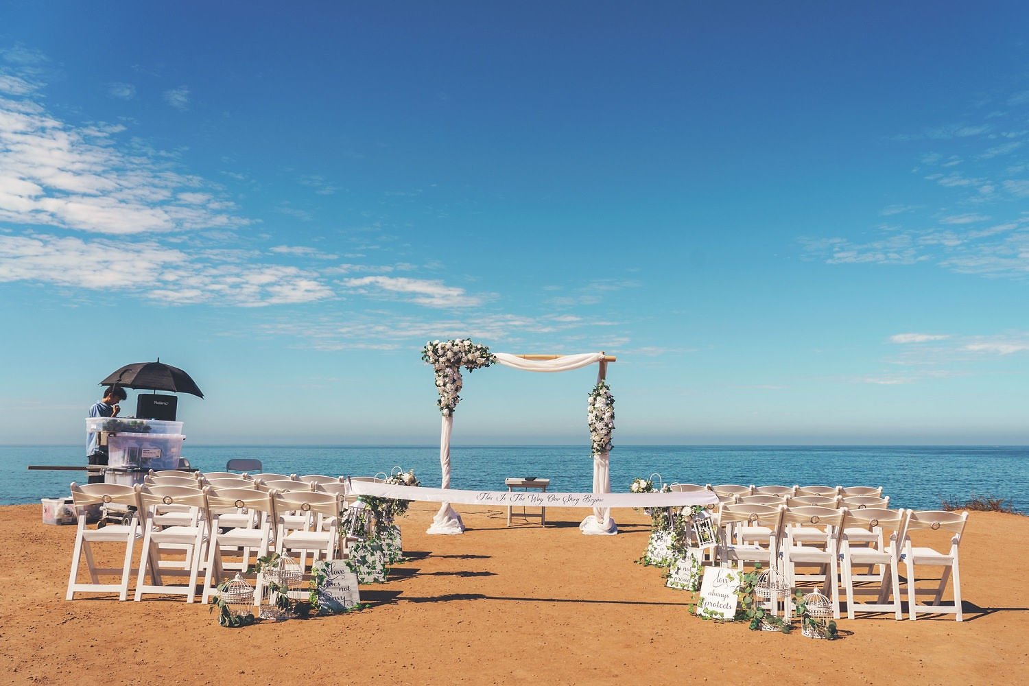 A beach wedding setup with white chairs arranged in two groups facing a floral arch over the ocean, decorated with white flowers and greenery. A person under a black umbrella is near a DJ station on the left, and the sky is clear with some clouds.