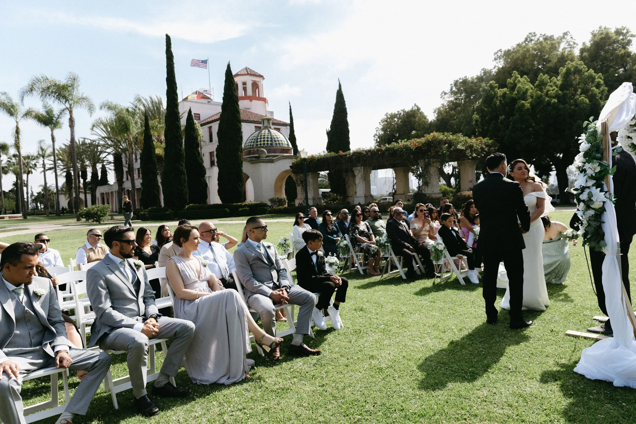 A wedding ceremony taking place outdoors on a grassy lawn with seated guests, a couple standing at the altar, and a white building with a dome and tall cypress trees in the background.